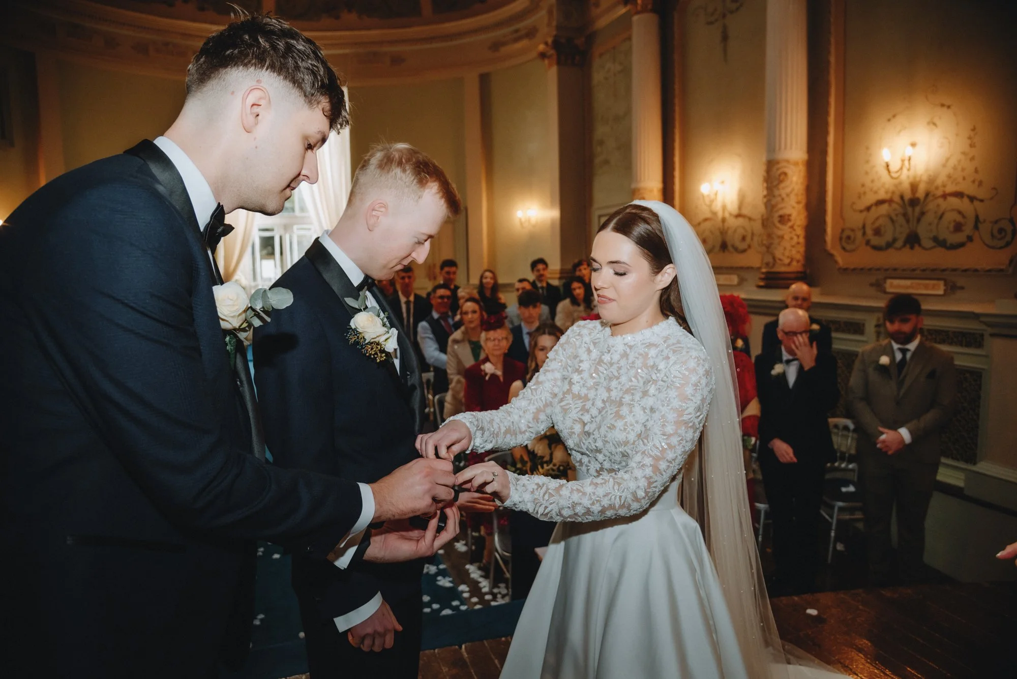 A bride and three grooms exchange rings during a wedding ceremony in an elegant room filled with seated guests.