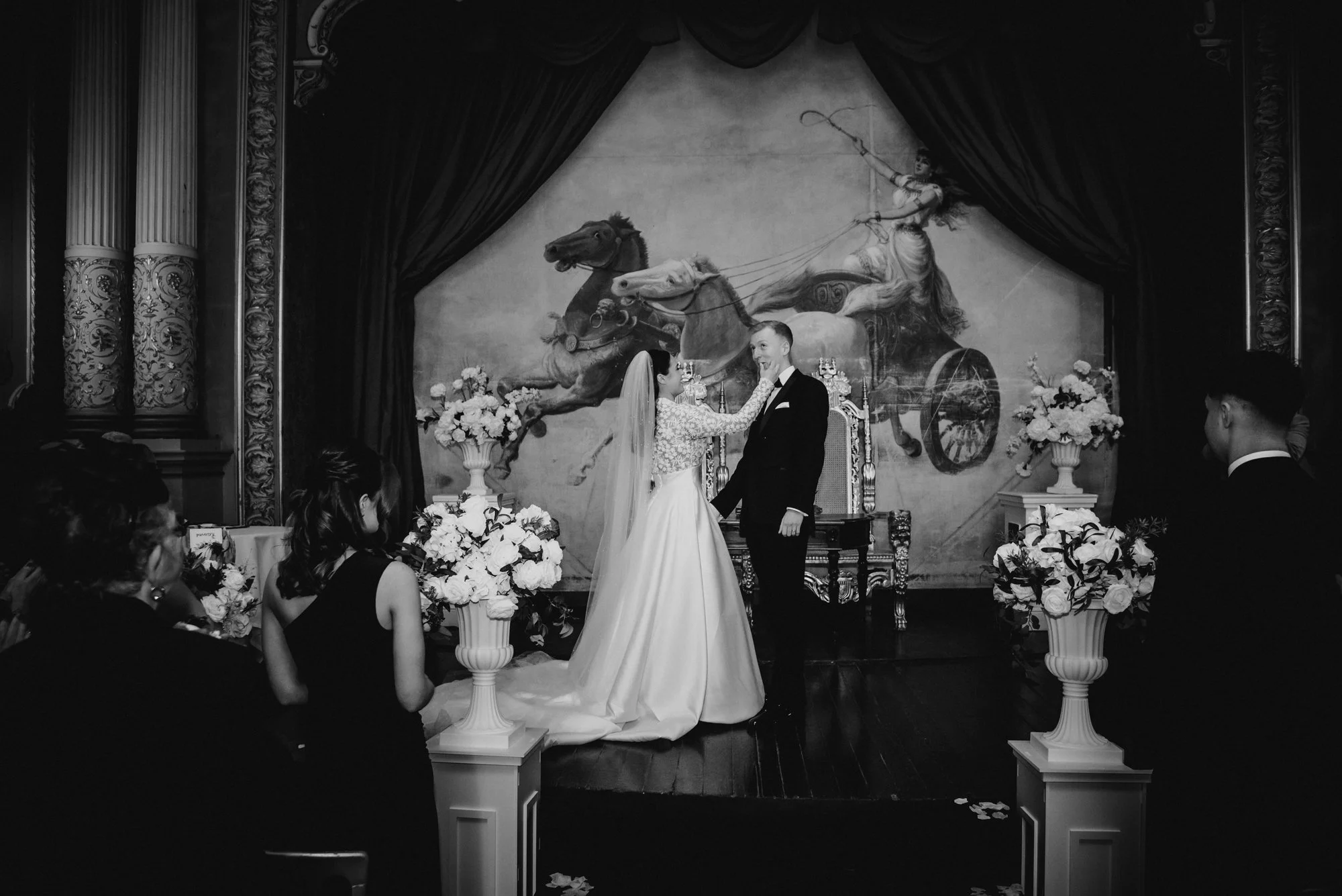 A black and white photo of a wedding ceremony with a bride and groom standing in front of an altar adorned with flowers, with a mural of a chariot drawn by horses in the background.