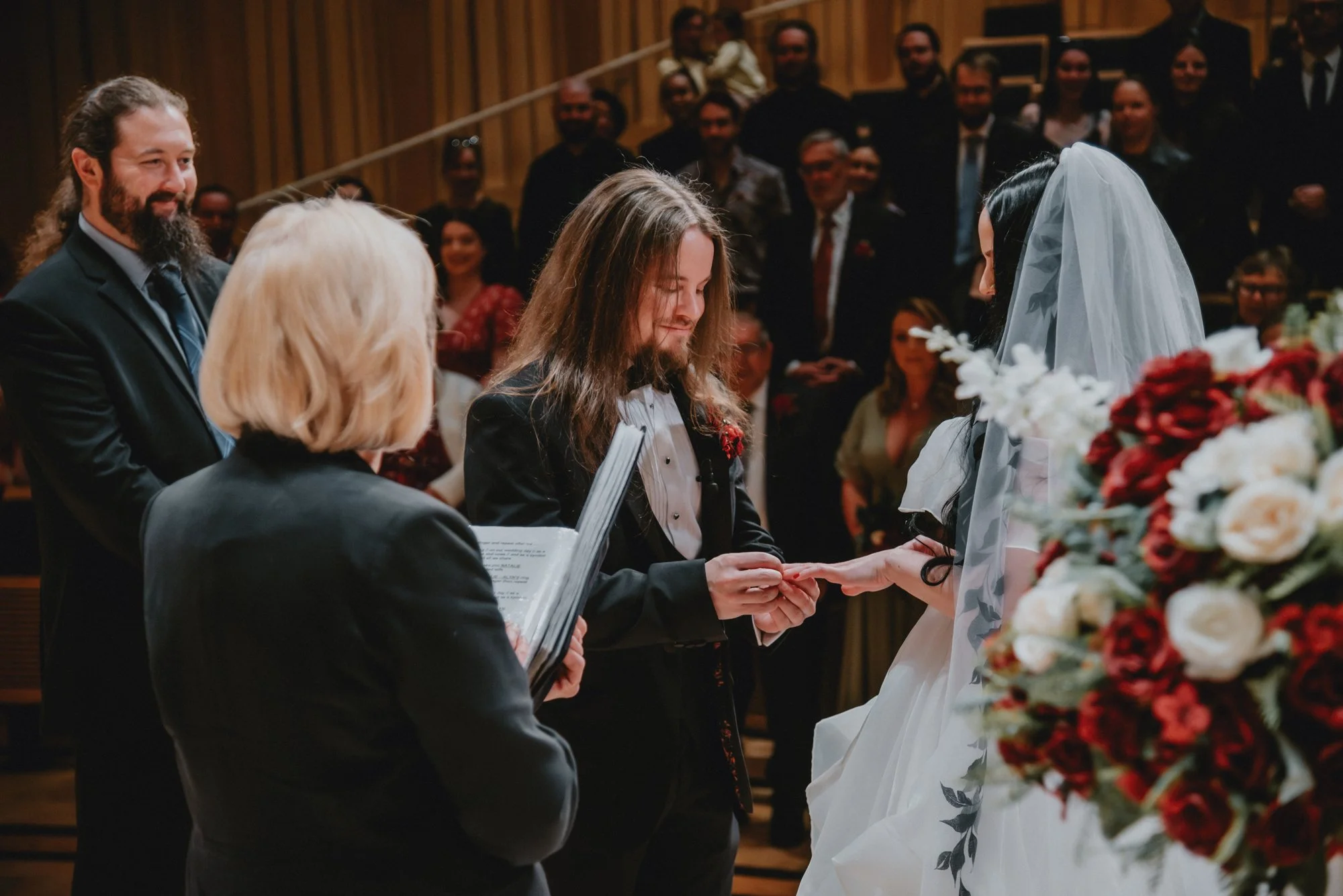 A wedding ceremony with a couple exchanging rings. The groom has long hair and a beard, and the bride is wearing a white veil. An officiant holds a book while the guests look on smiling.