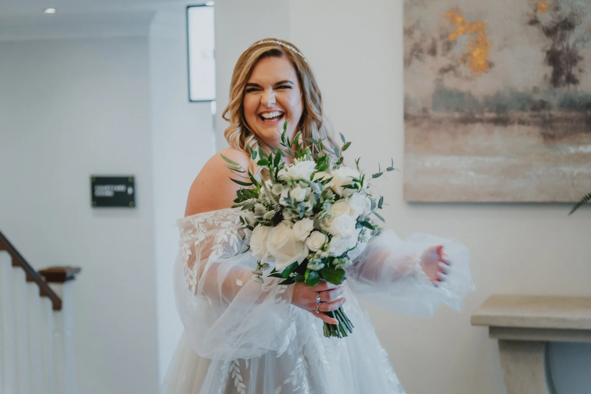 A happy bride in a white wedding dress holding a large bouquet of white roses and greenery, smiling brightly.