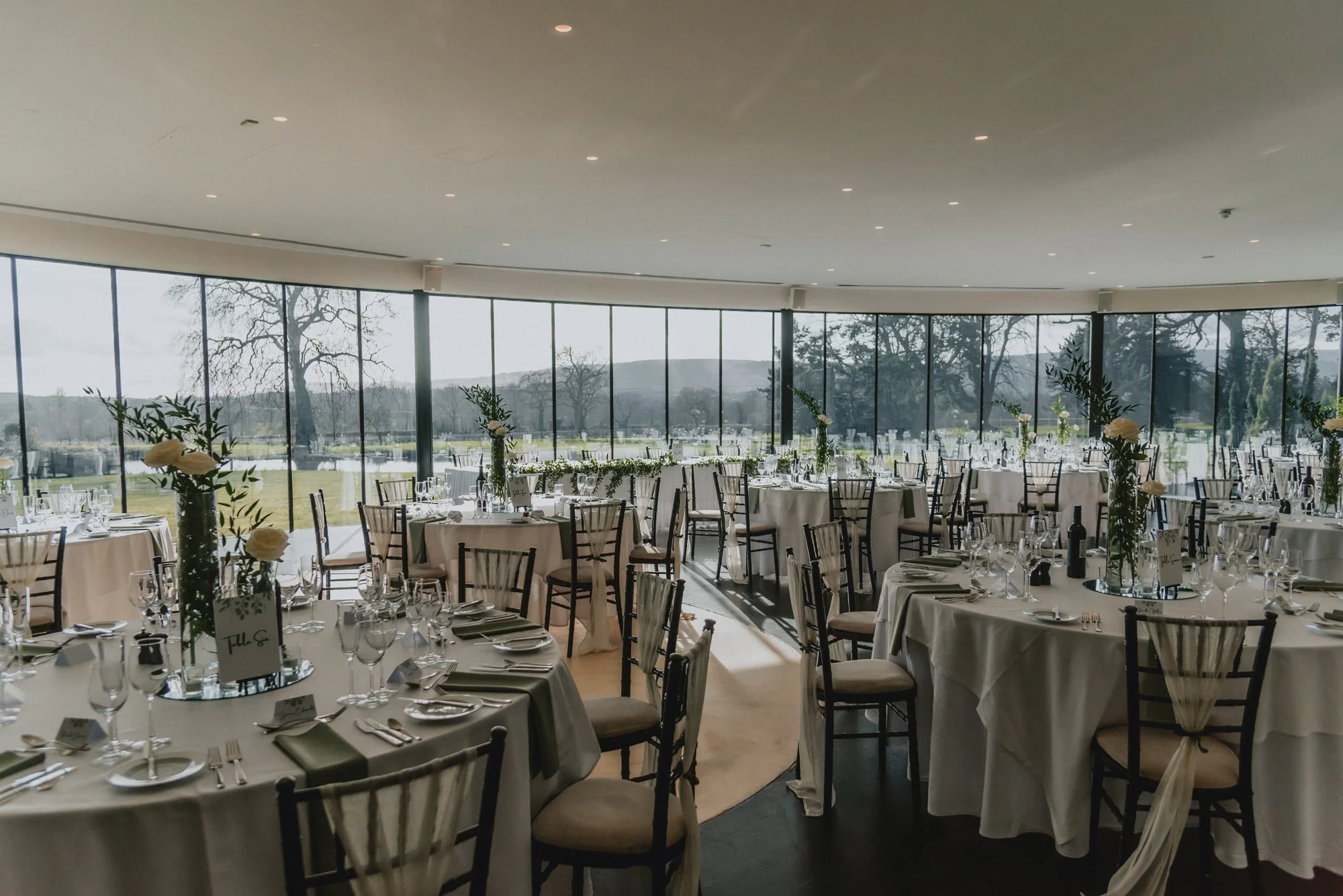 Elegant banquet hall with round tables covered in white tablecloths, decorated with tall floral centerpieces, set with glasses, silverware, and napkins, overlooking large windows with scenic outdoor landscape.