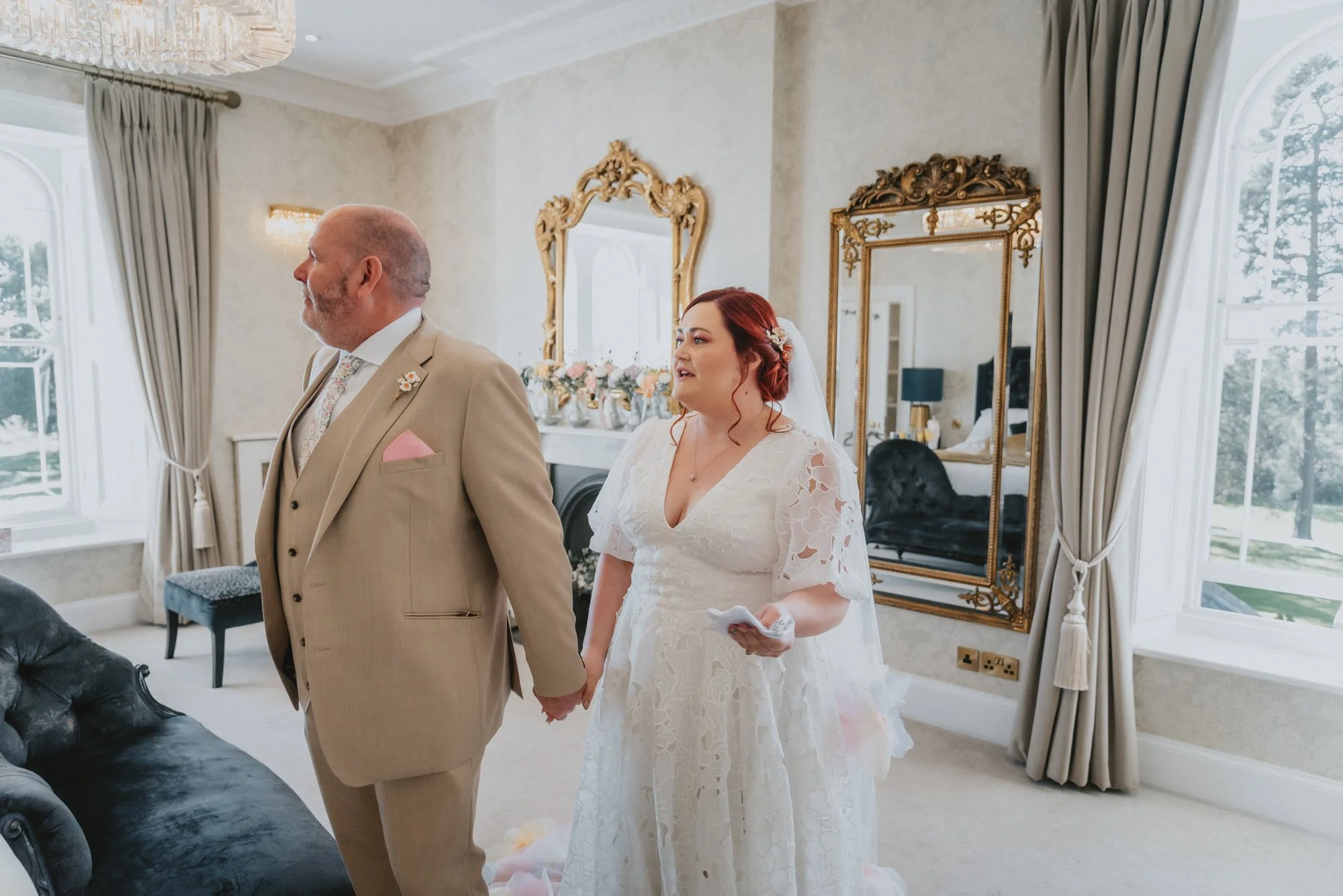 Bride and groom holding hands during wedding ceremony in elegant room with large windows, gold-framed mirrors, and floral decor.