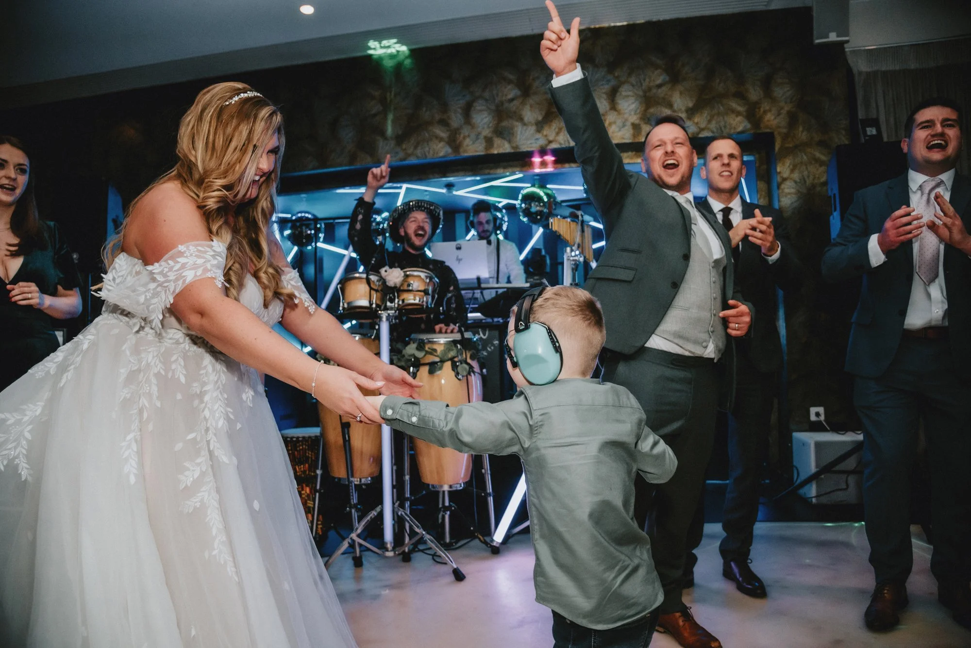 Wedding reception with a bride and young boy dancing, surrounded by guests, band members playing drums and keyboards in the background.