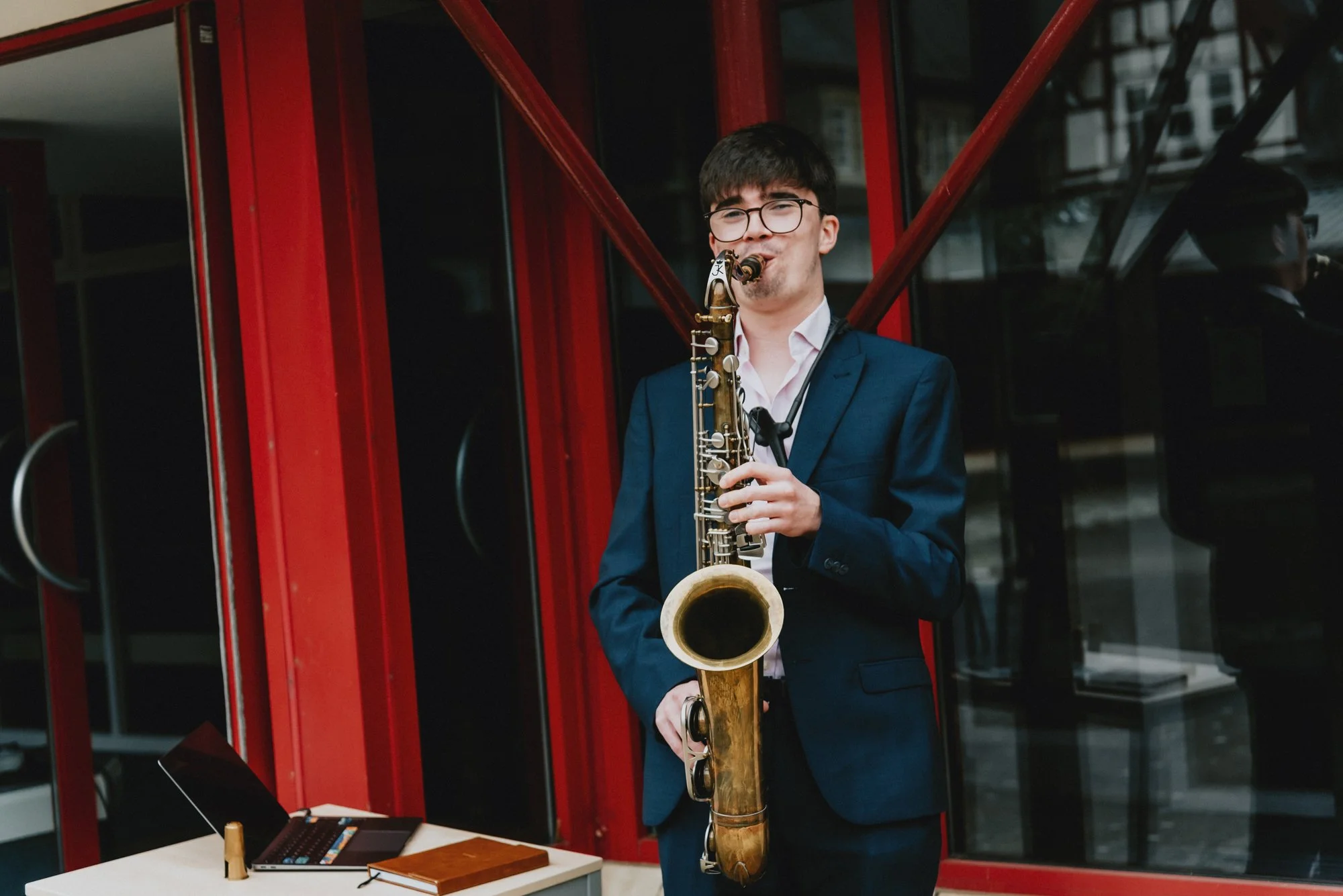 A young man in a navy suit and glasses playing a saxophone outdoors near a red and glass building.