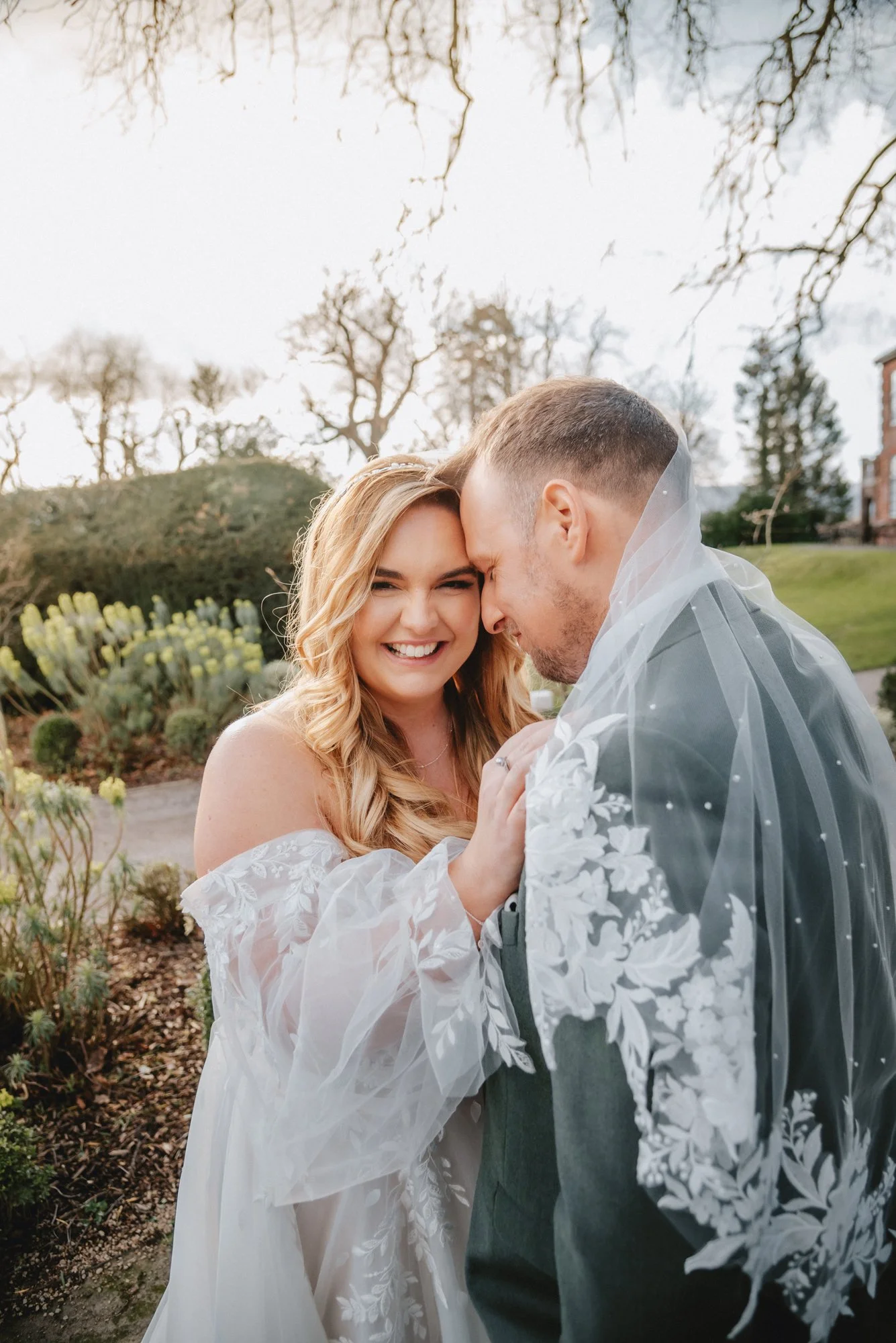 A joyful bride and groom on their wedding day, holding hands and smiling, outdoors with trees and greenery in the background.