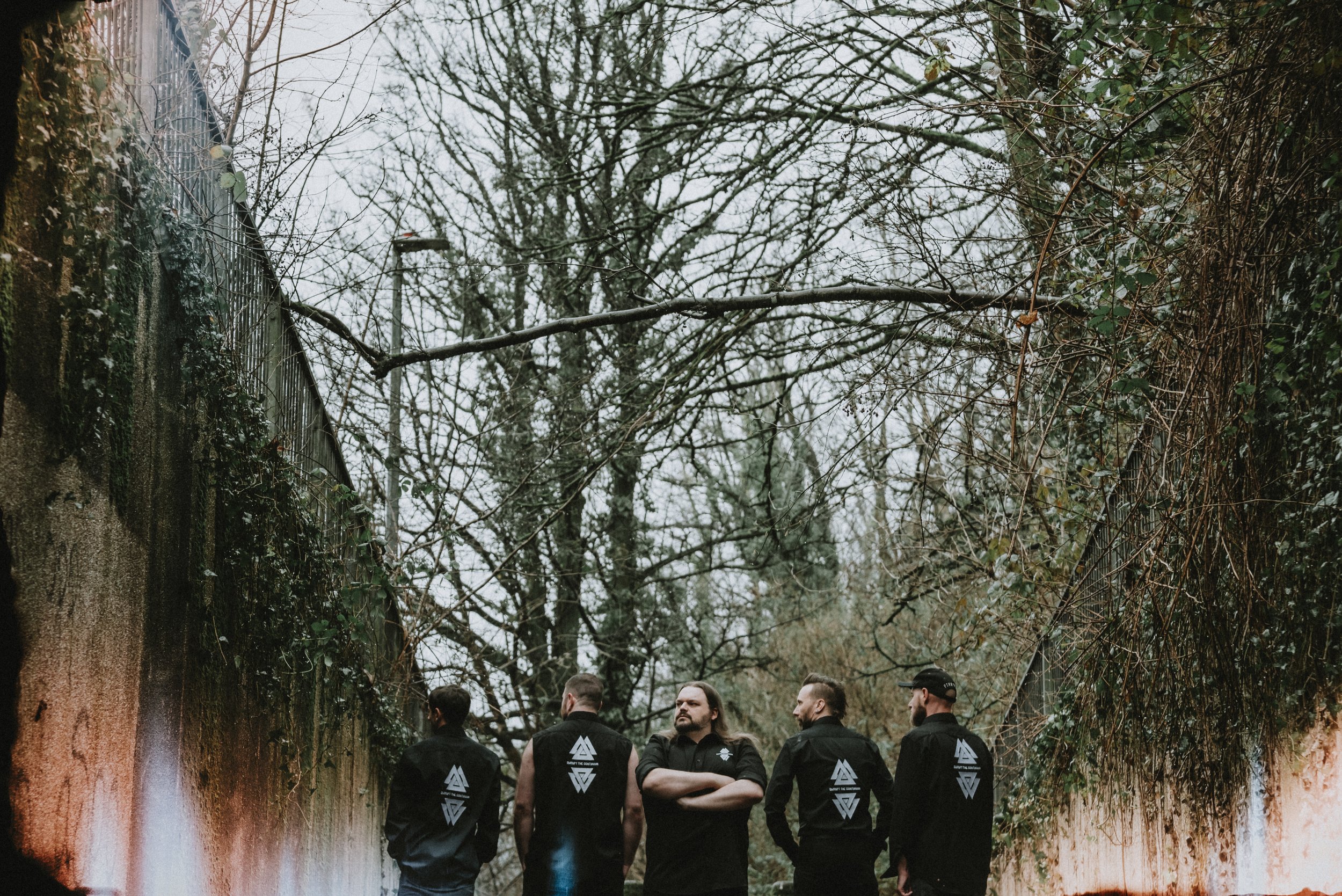 Five men with long hair or beards, wearing black jackets with white geometric logos on the back, standing outdoors among leafless trees and overgrown vines on a fence, in a cloudy, overcast setting.