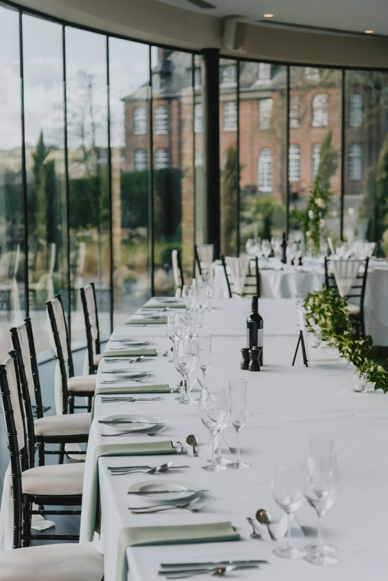 Elegant dining room with a long white table set for a formal occasion, featuring multiple wine glasses, silverware, and napkins, with large windows and a scenic view of a historic brick building outside.