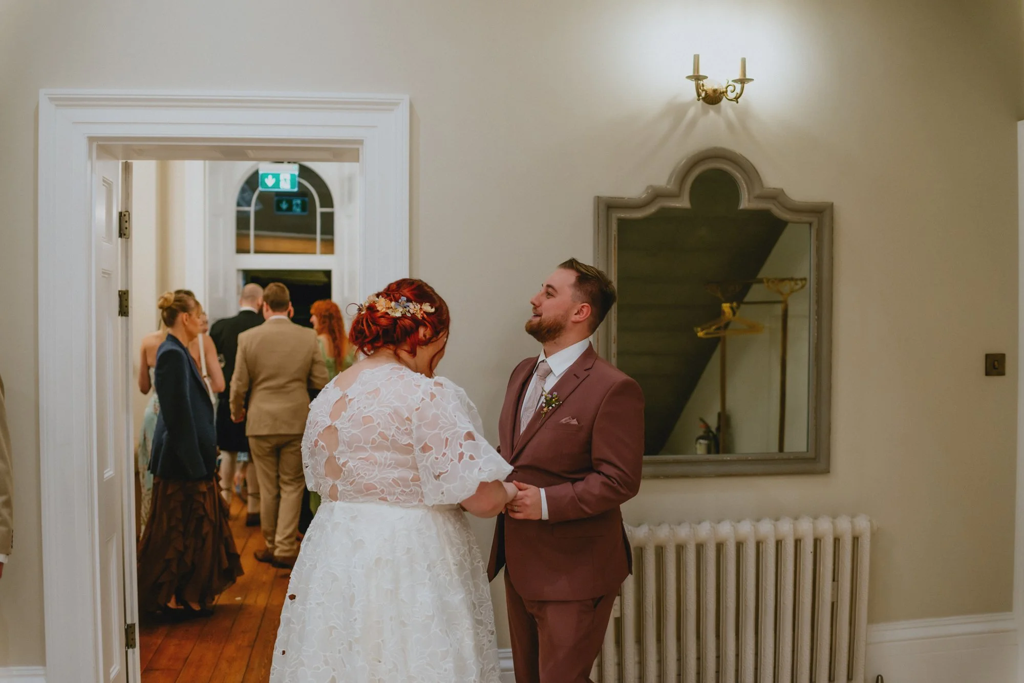 A bride and groom holding hands and smiling at each other during their wedding ceremony indoors, with wedding guests in the background.