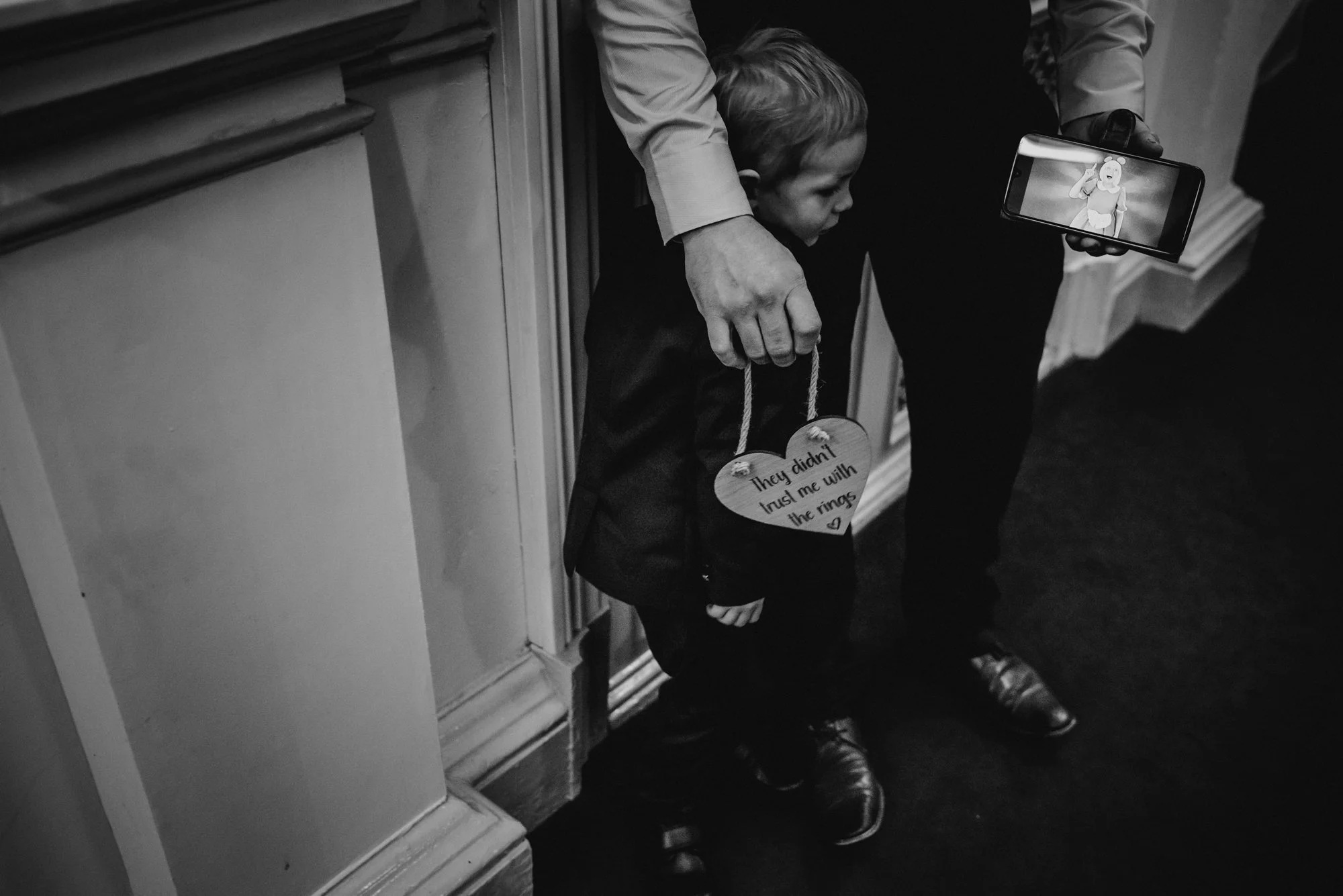 A young boy dressed in a suit, standing on a carpeted floor, holding a heart-shaped sign that reads 'They didn't trust me with the rings,' while a person behind him, partly visible, is holding a smartphone, taking a picture of him.