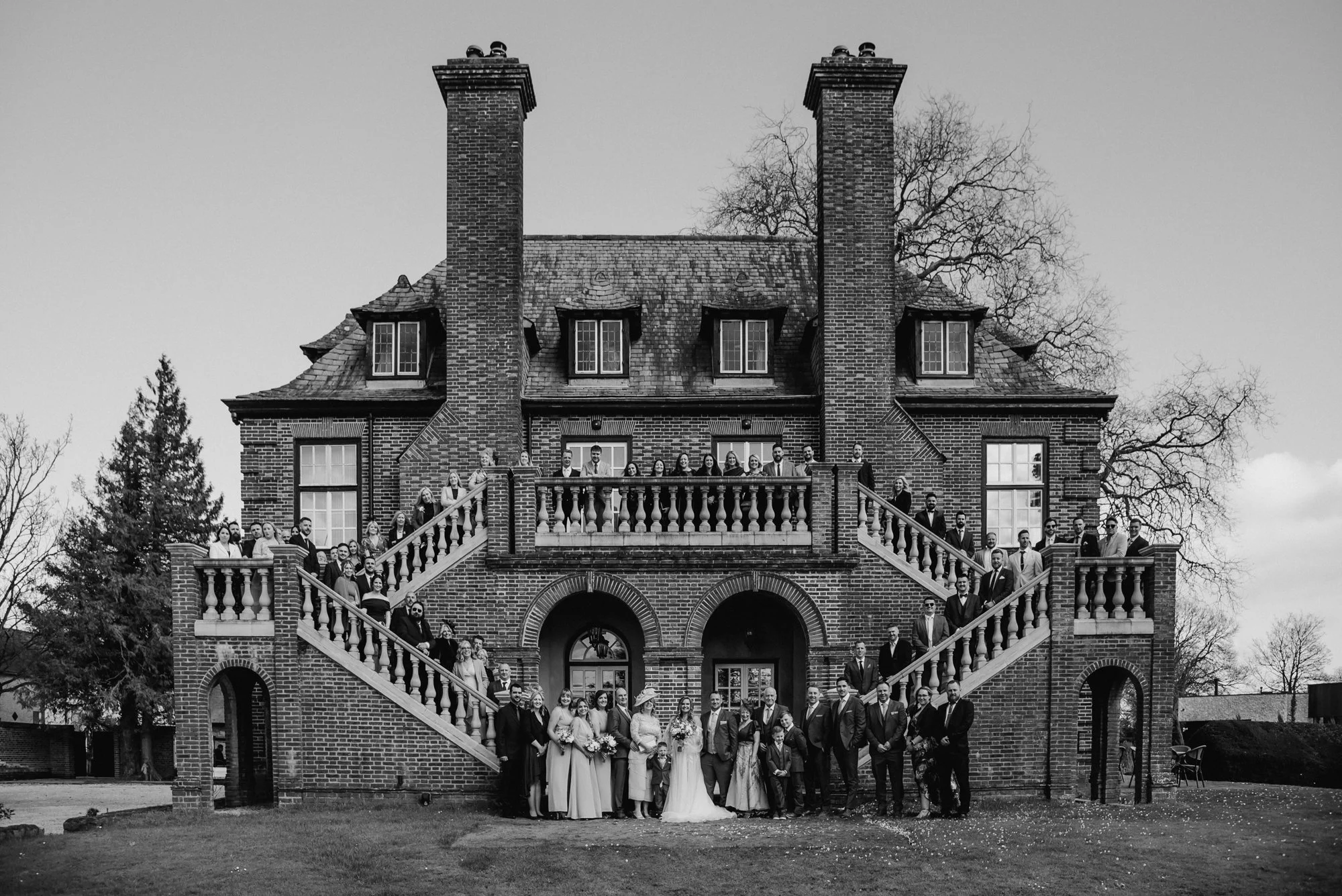 A large brick house with double staircases leading up to a balcony, filled with a group of people dressed in formal clothing, including a bride and groom at the center.