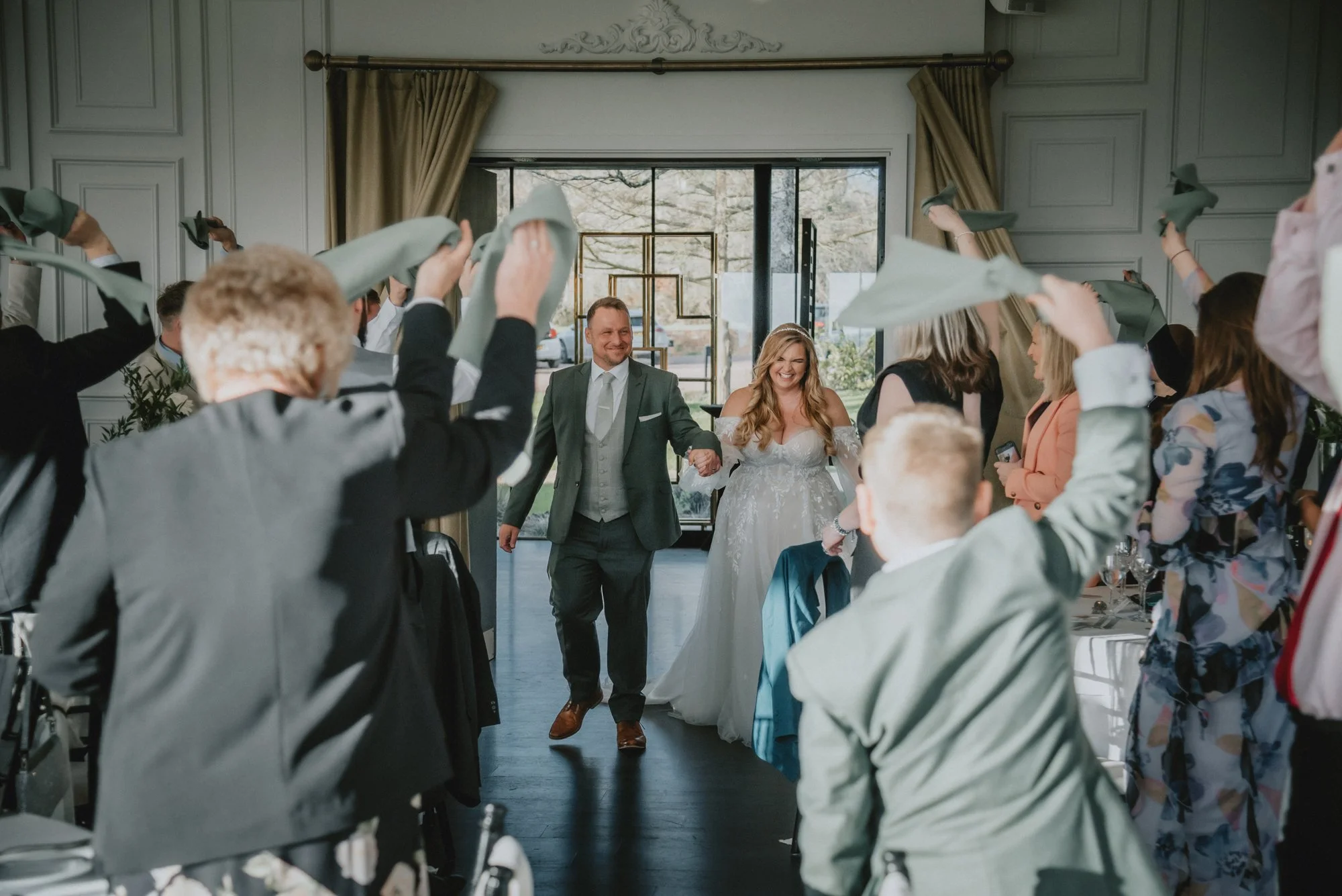 Bride and groom entering a wedding reception surrounded by guests waving napkins in celebration.