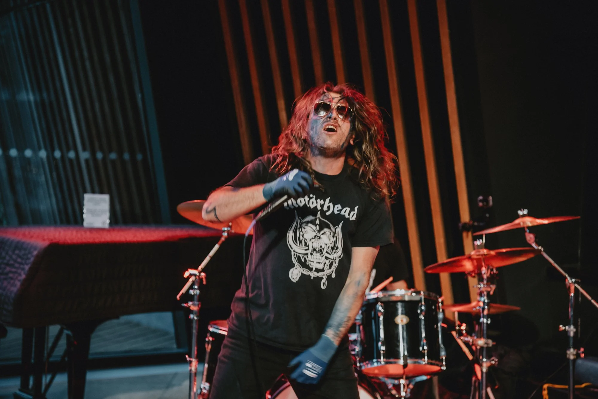 Musician with long hair, sunglasses, and tattoos performing on stage with a microphone, wearing a Motörhead t-shirt, in front of drums and a piano.