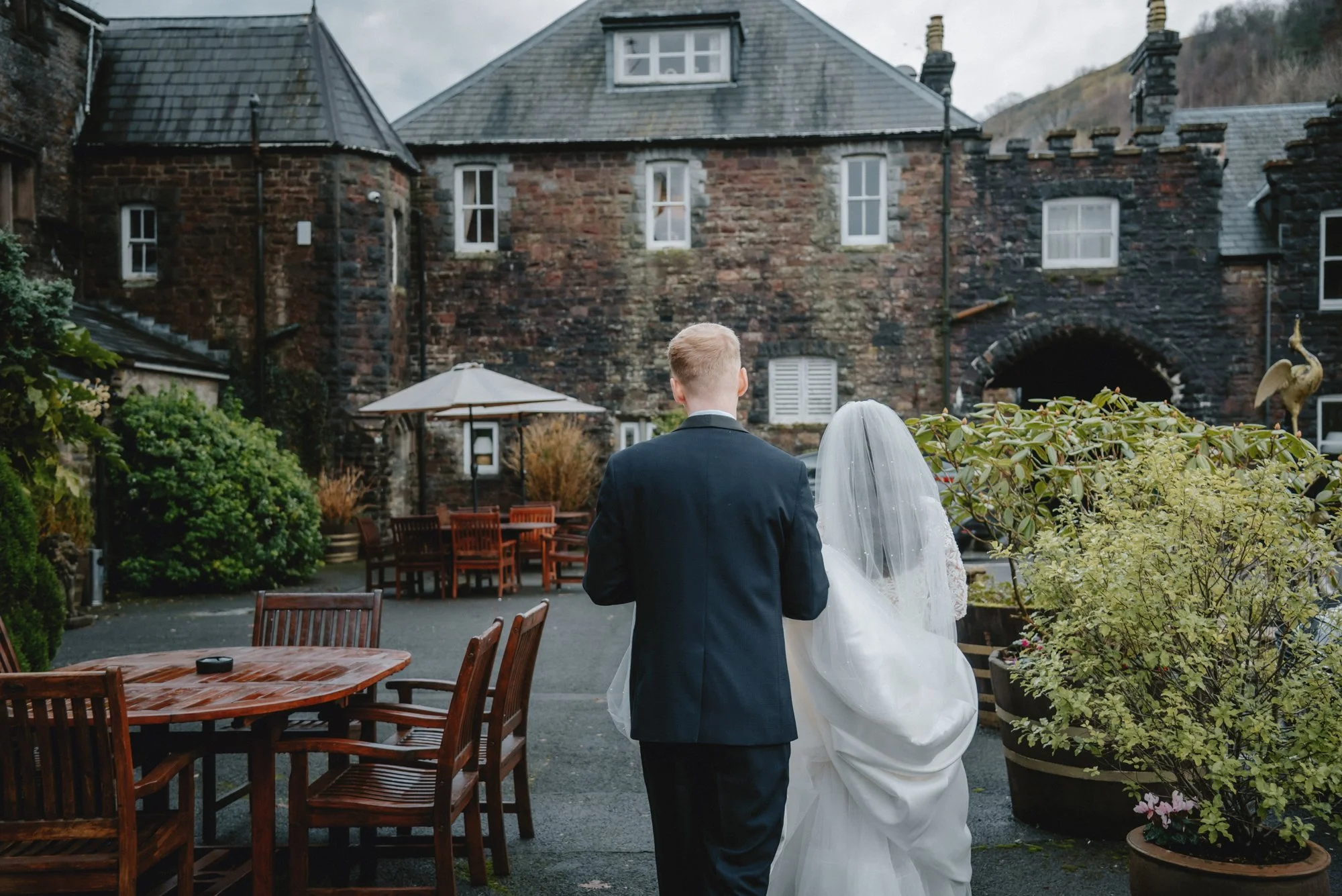 A bride and groom walking away from the camera in an outdoor venue with rustic brick buildings, wooden tables and chairs, greenery, and sky.