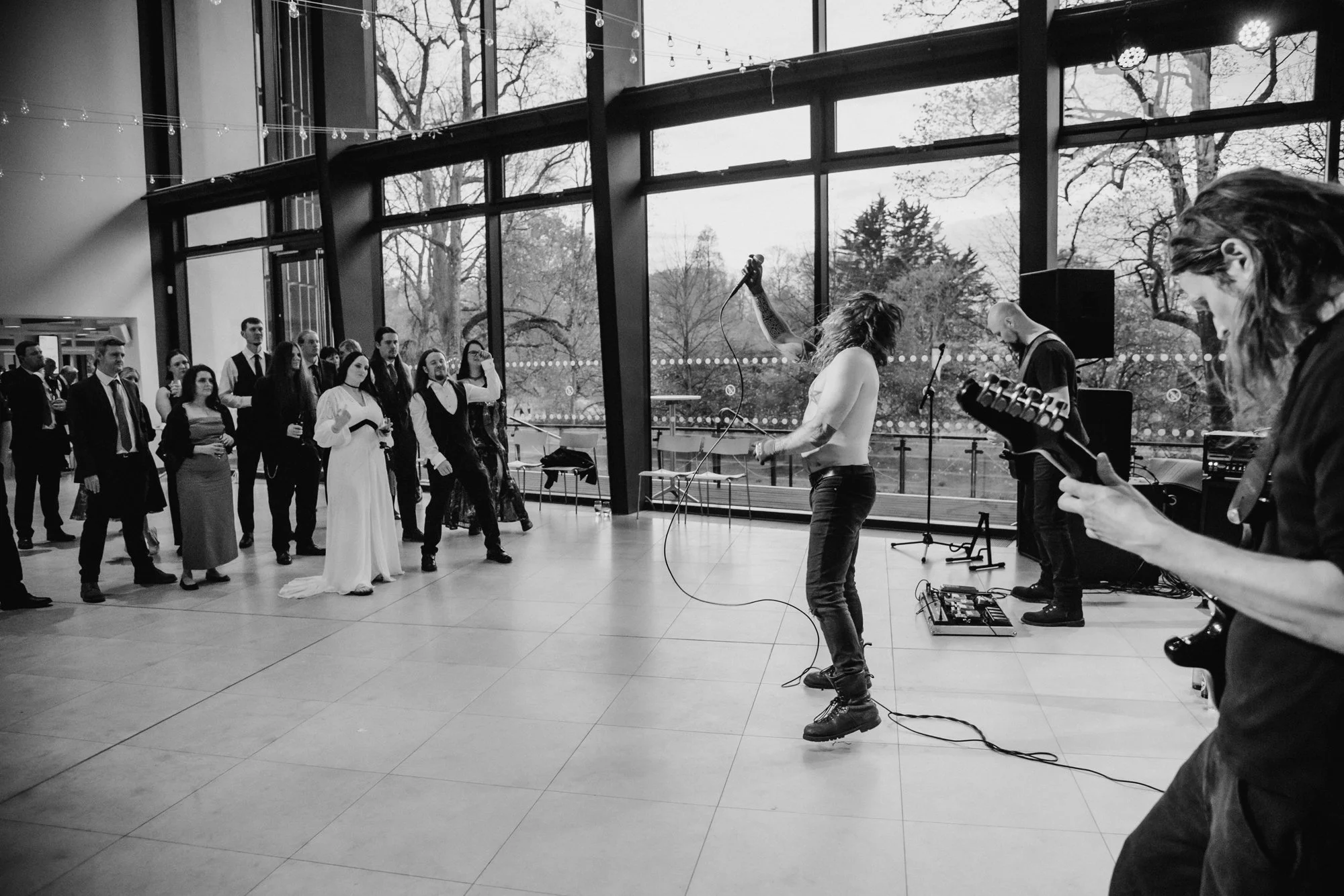 A band performs live music at an event with people dancing in front of large windows showing trees outside. The band features a shirtless guitarist, a singer holding a microphone, and another member playing an electric guitar. The crowd includes men 