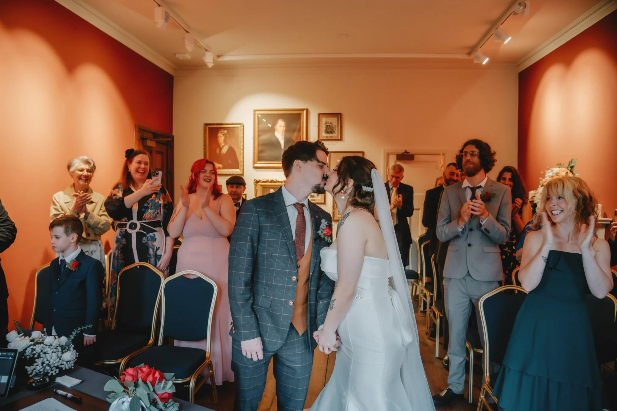 A wedding ceremony with a couple kissing in the center, surrounded by seated and standing guests smiling and clapping in a decorated room with framed portraits on the wall.