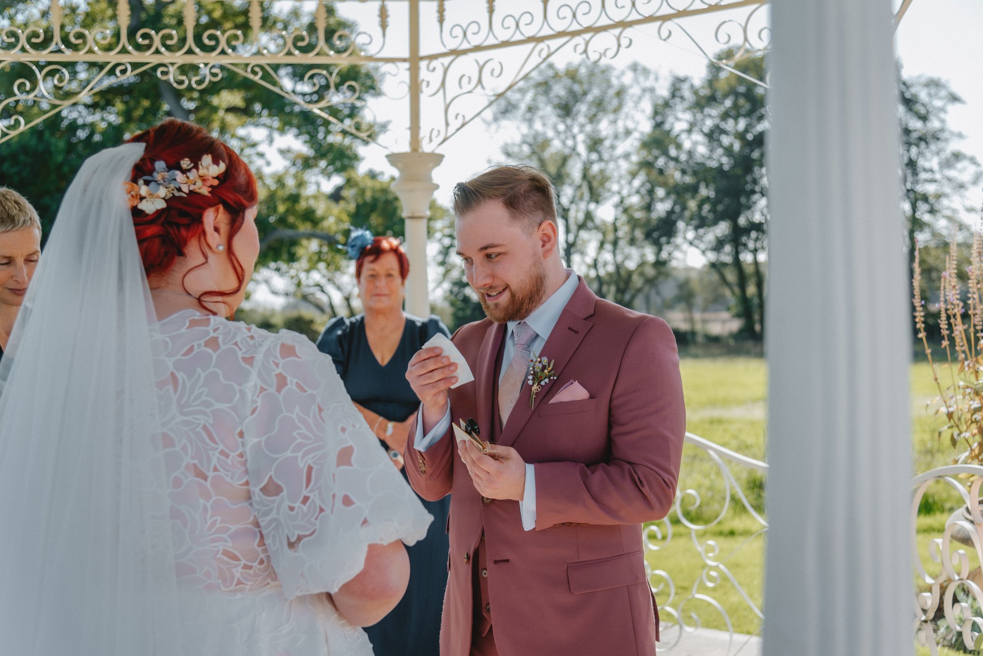 A wedding ceremony outdoors with a bride and groom exchanging vows. The bride has red hair with floral hair accessories and a lace veil. The groom has a beard and is wearing a mauve suit. There are women in the background, one with short blonde hair 