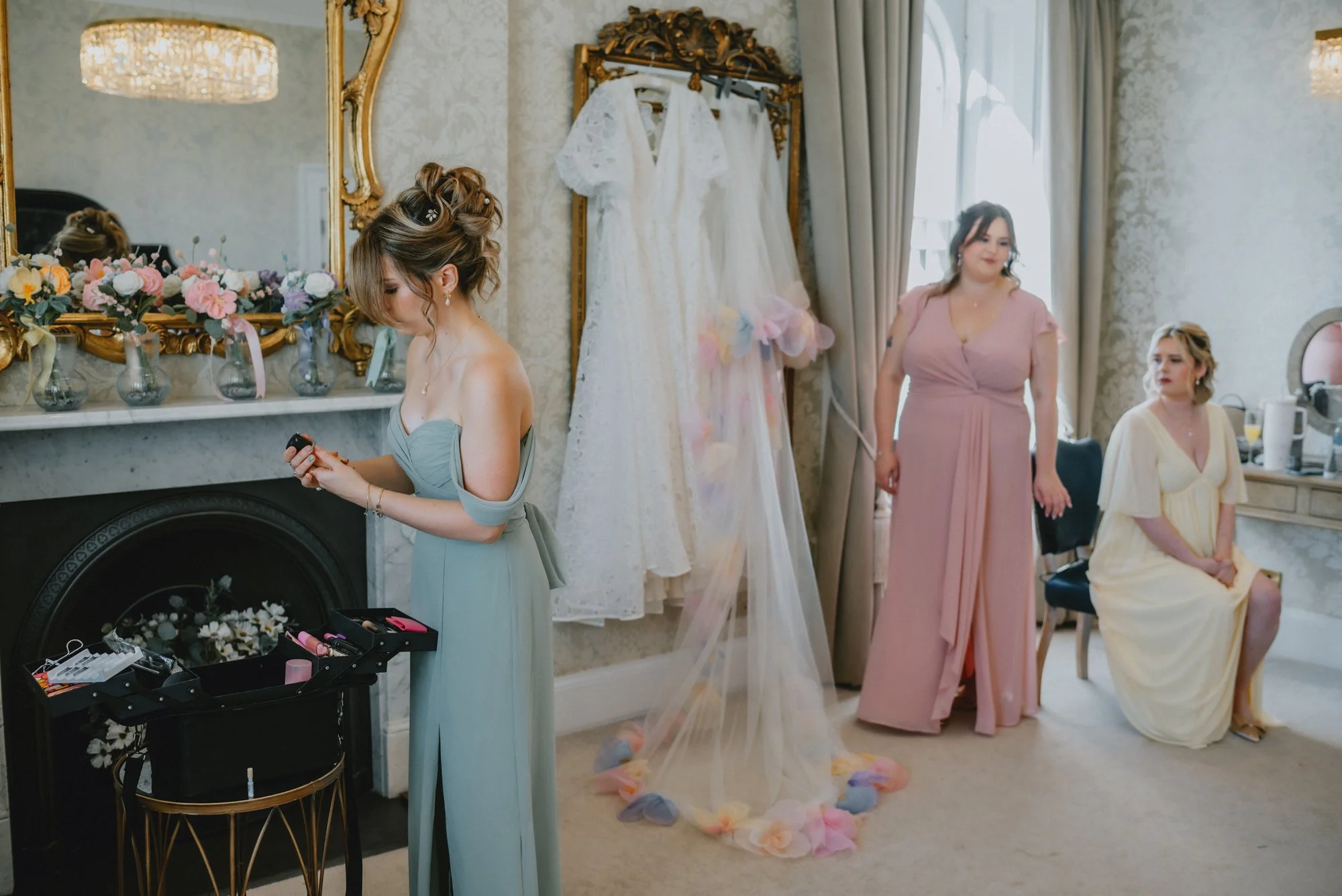 Three women getting ready in a bridal suite. One woman is in a silvery-gray dress, another in a pink dress, and the third in a yellow dress. A wedding dress is hanging on a golden ornate frame. The room is decorated with flower vases and a large mirr