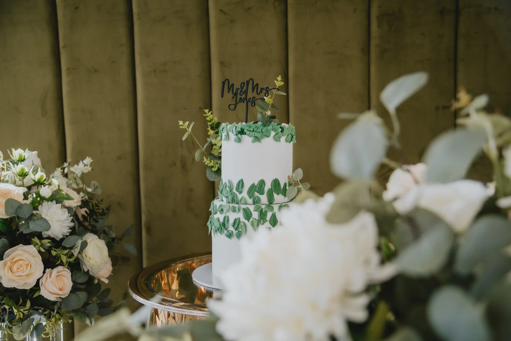 A white wedding cake decorated with green leaves and rosettes on top, placed on a reflective silver table, surrounded by flower arrangements, with a wooden background.