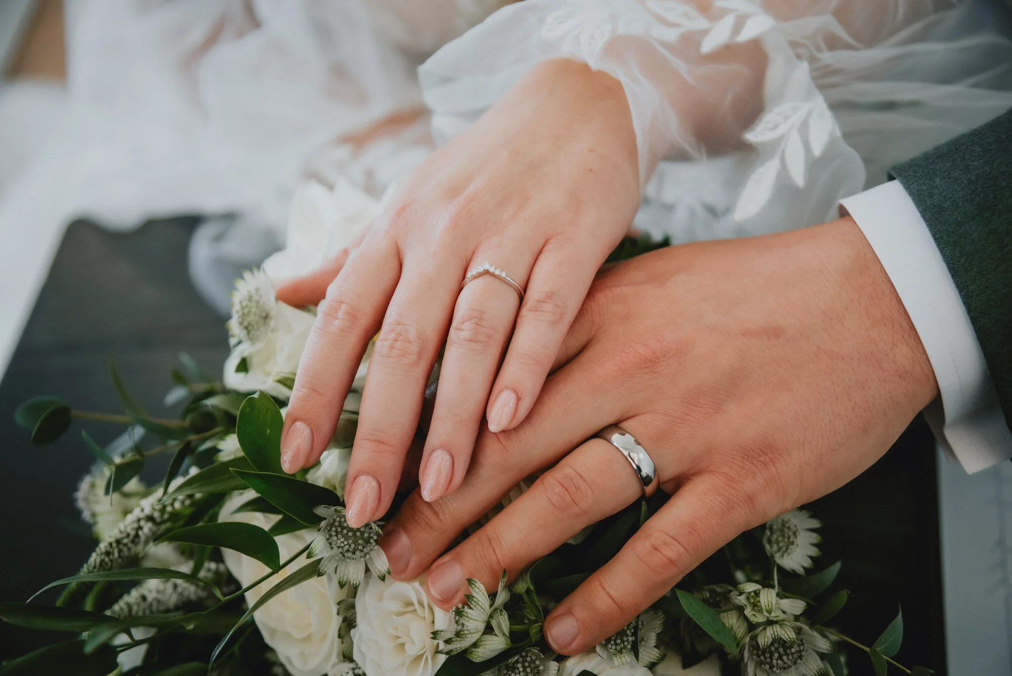 Close-up of a newlywed couple's hands with wedding rings, resting on a bouquet of white roses and greenery.