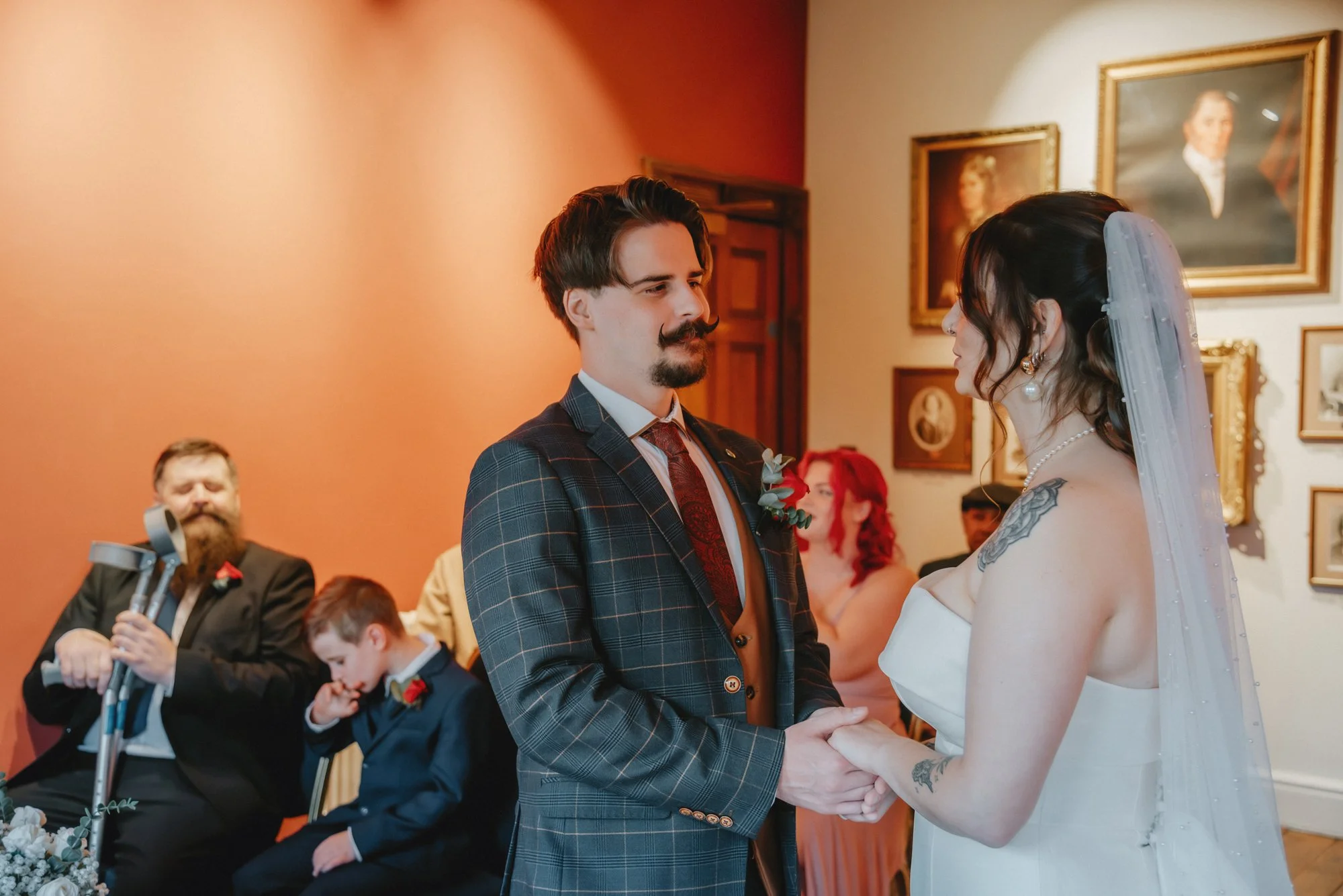 A couple is holding hands and looking into each other's eyes during a wedding ceremony. The groom has dark hair, a mustache, and a beard, and is wearing a checkered suit with a boutonniere. The bride has long dark hair, a veil, and a tattoo on her ar