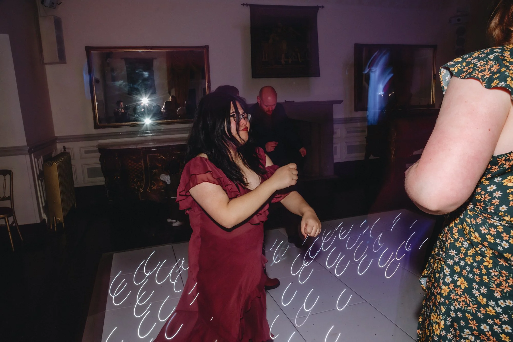 A woman in a maroon dress dancing on a lit dance floor indoors, with three other people in the background, and a woman in a floral dress partially visible on the right side of the image.