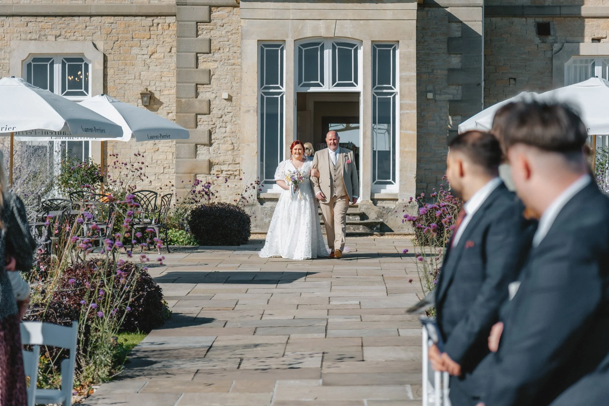 Bride walking down the aisle with her father at an outdoor wedding ceremony, with guests seated on either side and a stone building in the background.