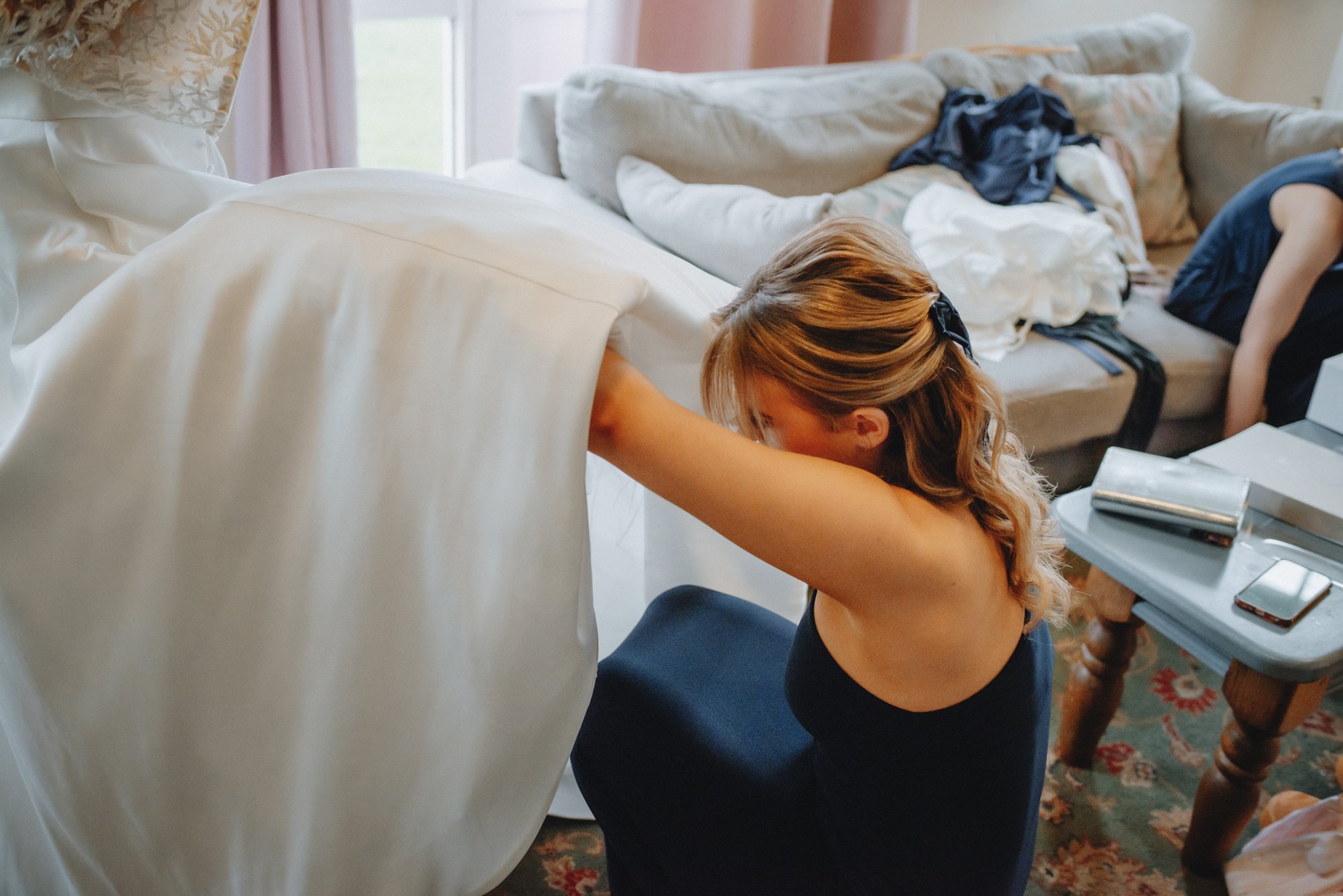 A woman with light brown, wavy hair tied with a black hair clip, wearing a dark blue dress, is kneeling on a patterned rug and stretching her arms upward, appearing to help move a white piece of furniture in a cluttered living room. In the background