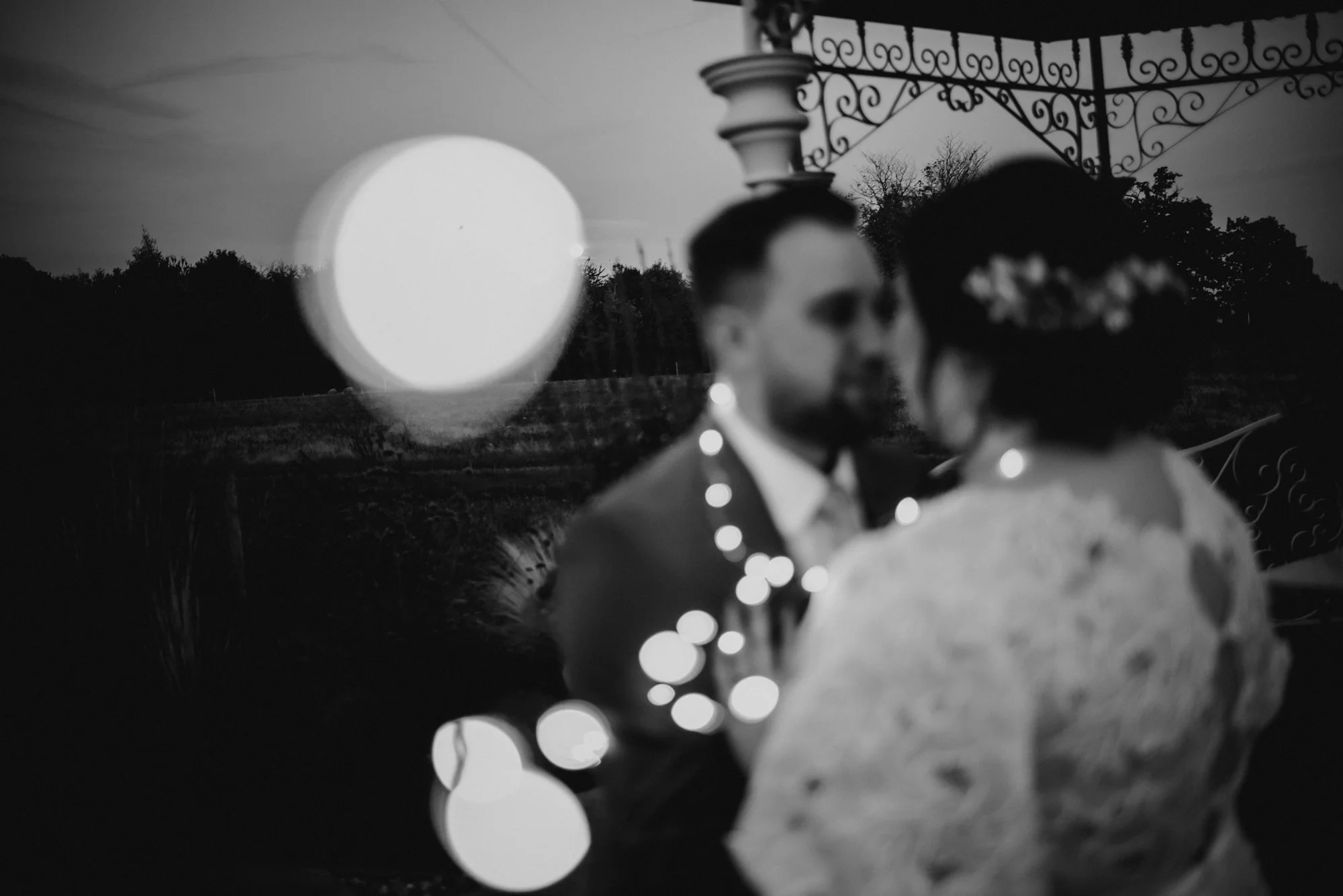 A black and white photo of a couple in wedding attire, with the bride wearing a lace dress and a floral headpiece, and the groom in a suit, standing close outdoors with trees and wrought iron structures in the background. There is a large out-of-focu