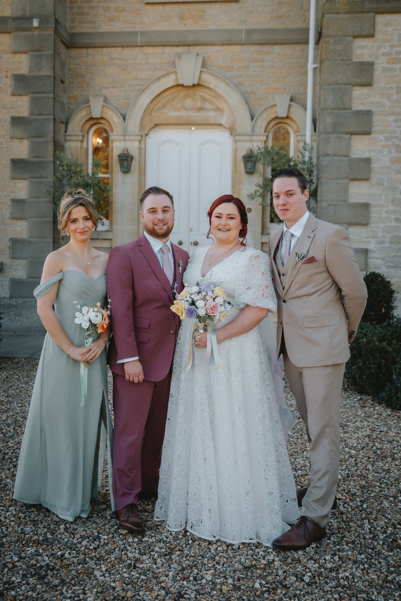 A wedding group photo of four people in front of a stone building, with the bride holding a bouquet and the groom in a burgundy suit, flanked by a bridesmaid in a gray dress and a groomsman in a beige suit.