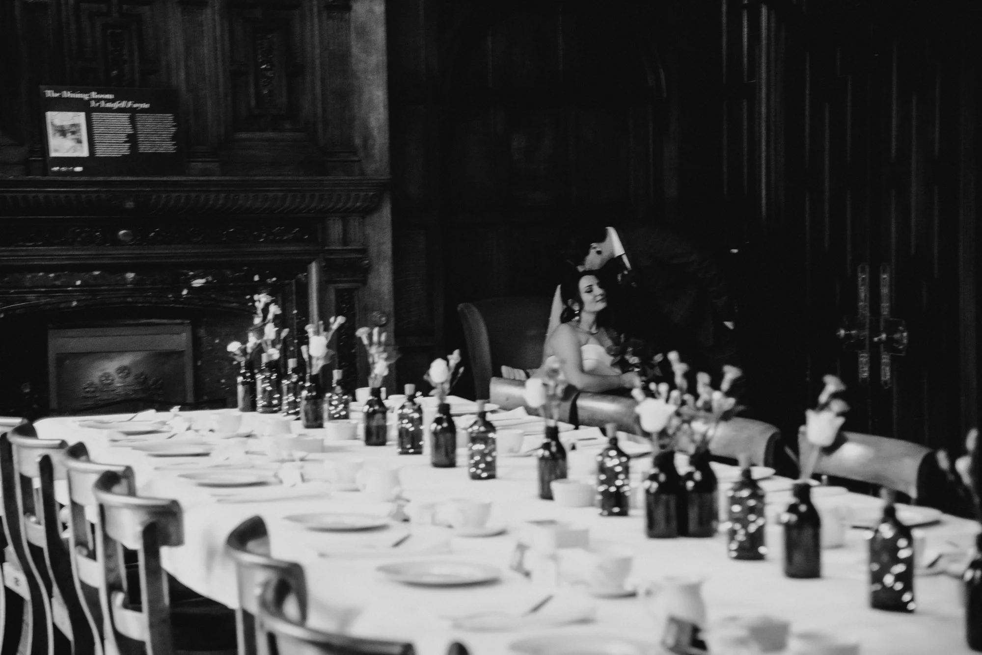 A woman in a wedding dress sitting in a chair at a decorated dining table with floral arrangements and bottles, in a dimly lit, elegant room.