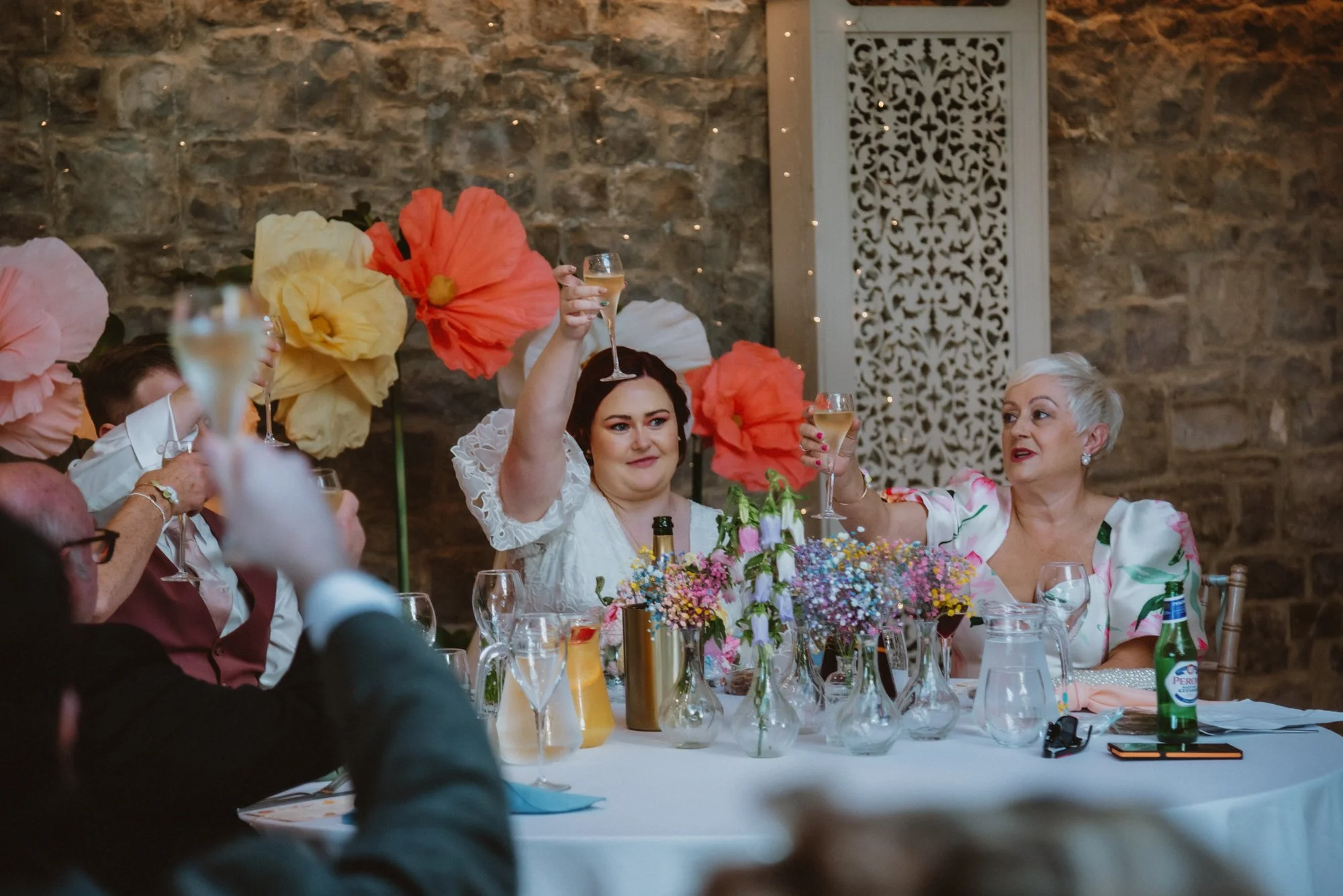 A group of women at a celebration raising glasses for a toast, with large colorful paper flowers and a decorated table with vases and flowers.