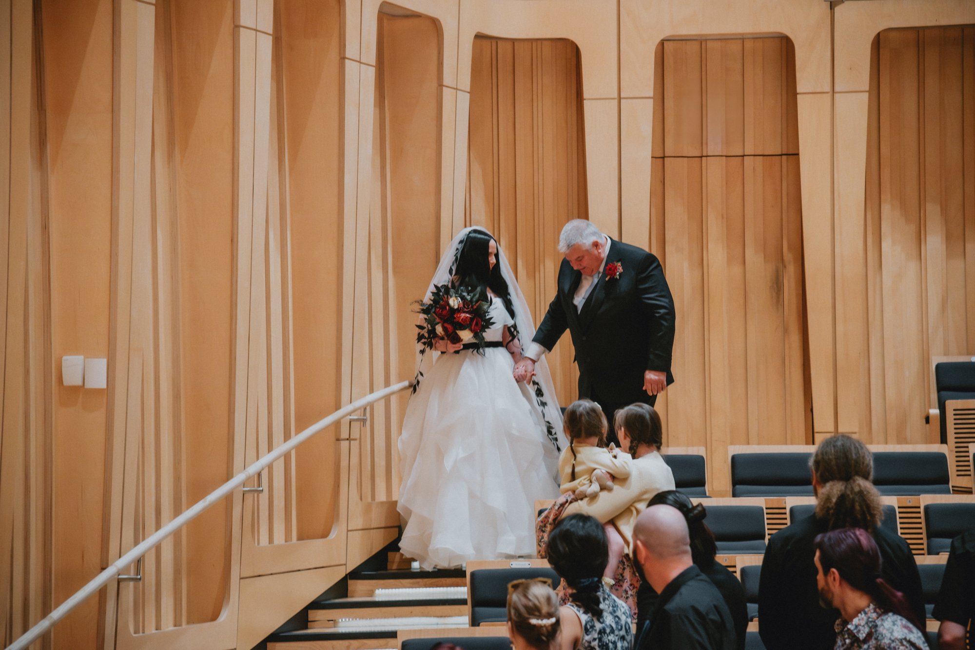 A bride and an older man, possibly her father, exchanging a handshake or a moment of blessing at a wedding ceremony. The bride is holding a bouquet and wearing a white gown with a veil, standing at the top of a small set of wooden stairs in an audito