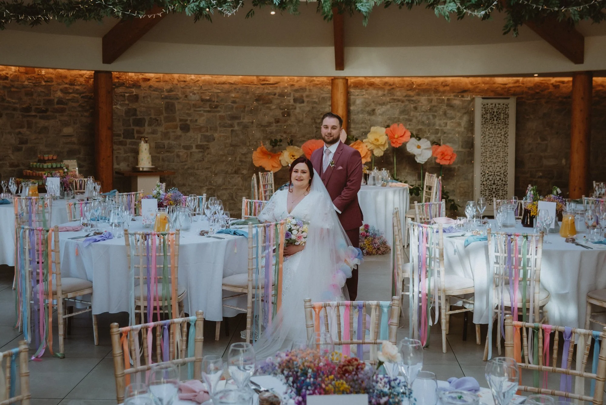 Bride and groom at their wedding reception, standing behind decorated tables with floral arrangements, in a venue with exposed brick wall, wooden beams, and colorful ribbons on chairs.