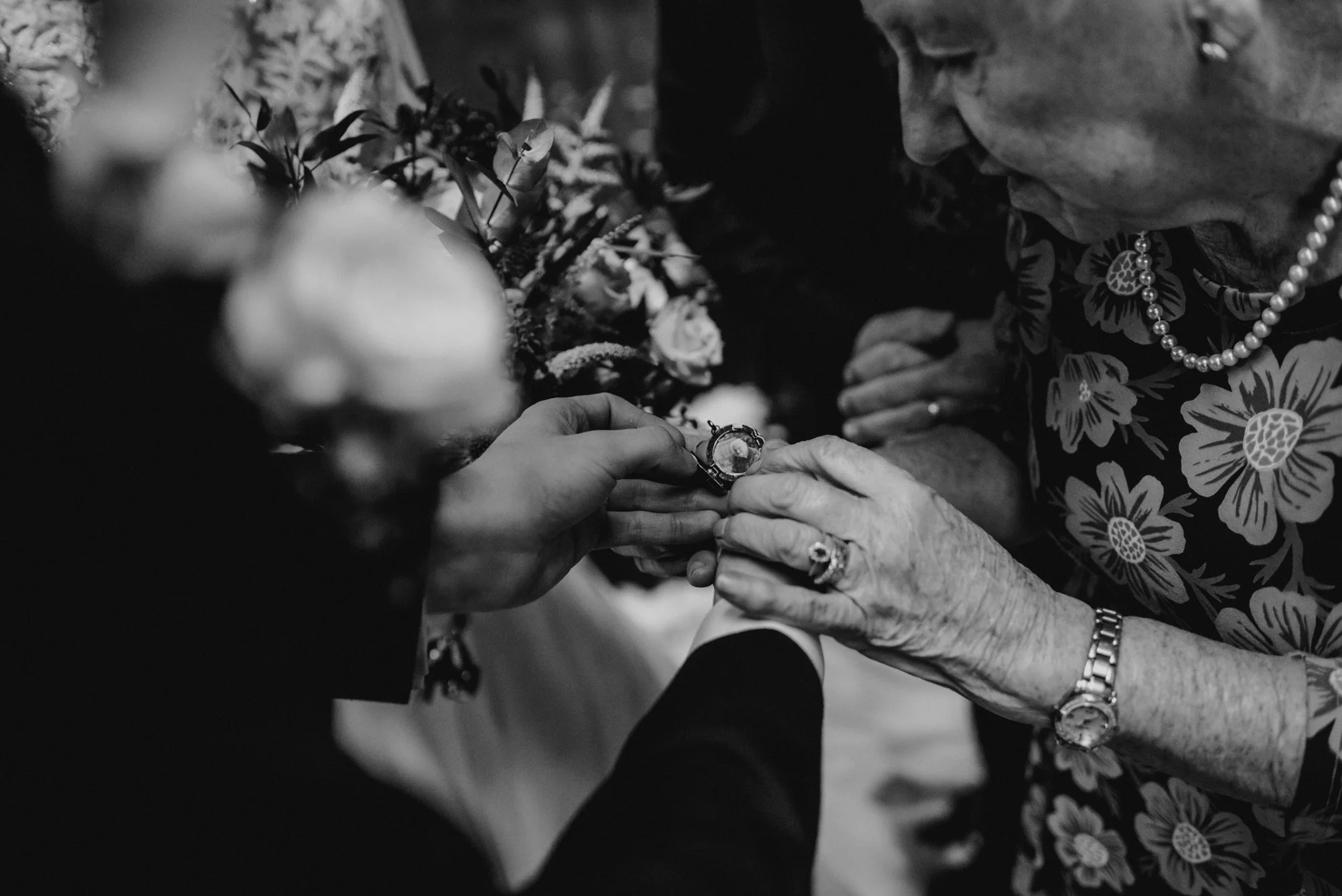 A black and white photo of an elderly woman with a pearl necklace and floral dress, receiving a ring from a younger person, with floral arrangements in the background.