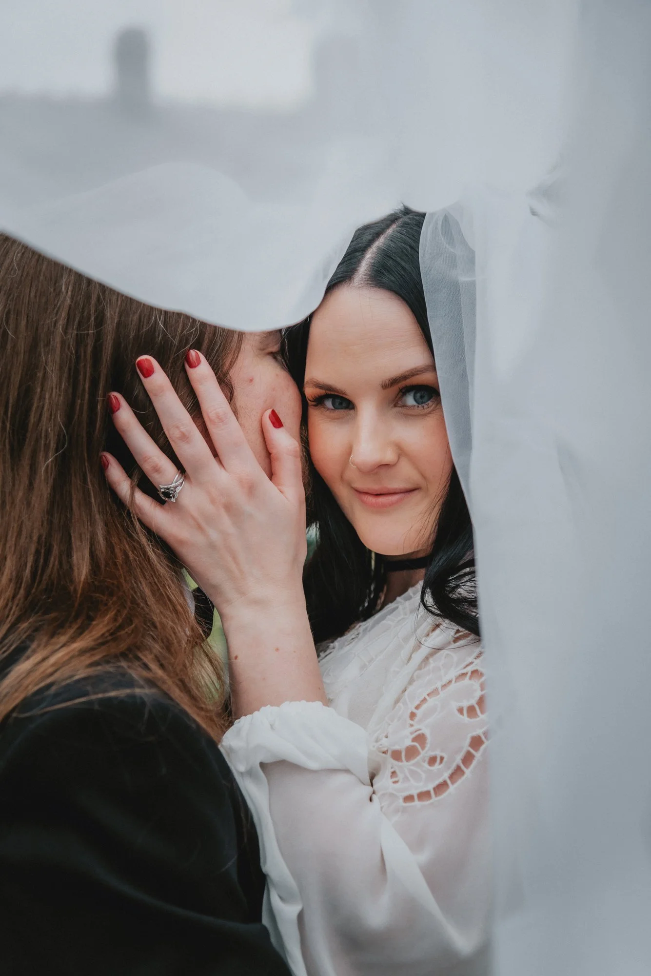 Two women sharing an intimate moment behind a semi-sheer white curtain, one woman with dark hair and blue eyes and the other with red hair and a wedding ring.