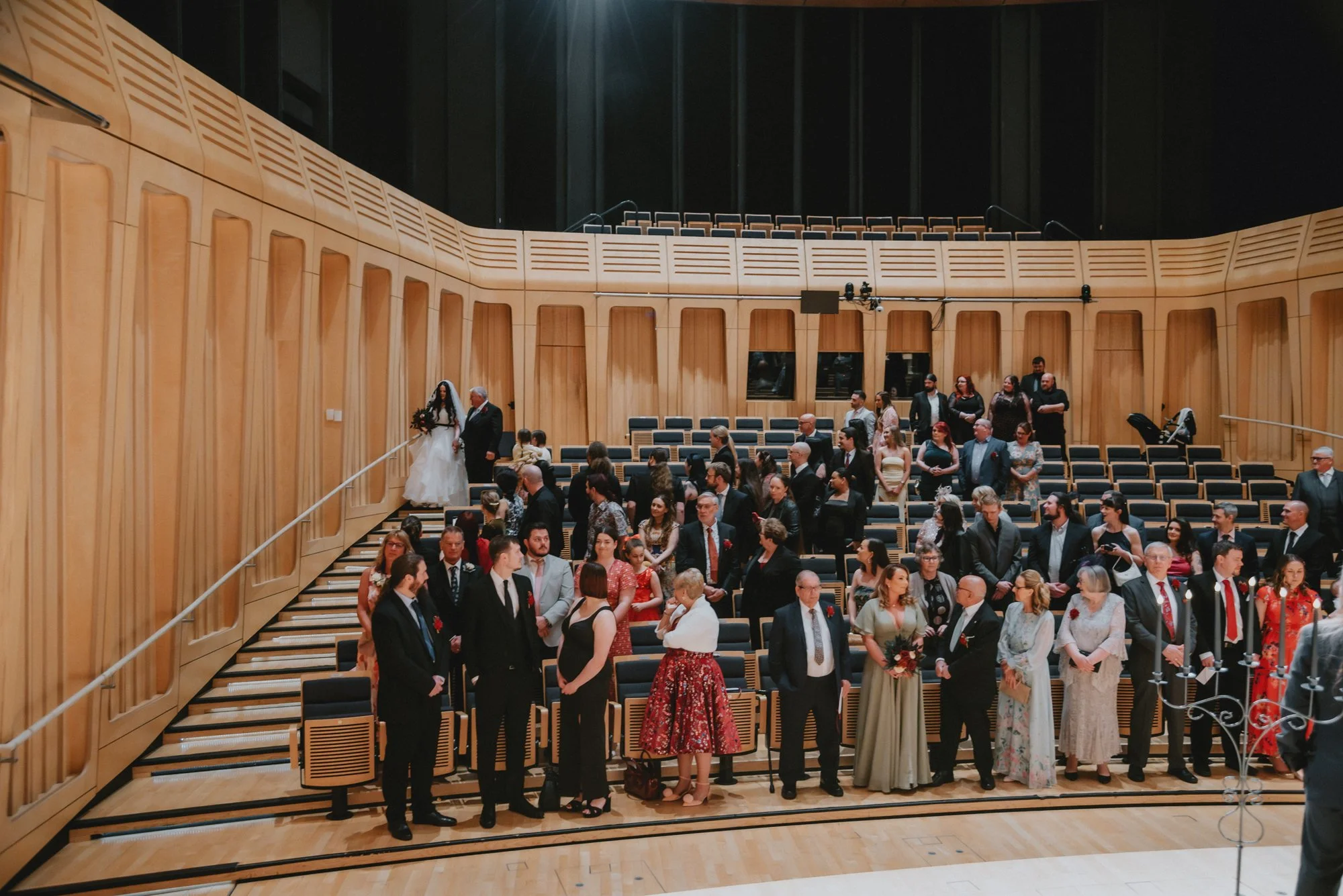 People attending a wedding inside a concert hall or auditorium with wooden paneling on the walls, some people standing and others sitting, a bride and groom walking down stairs, and a few individuals taking photos.