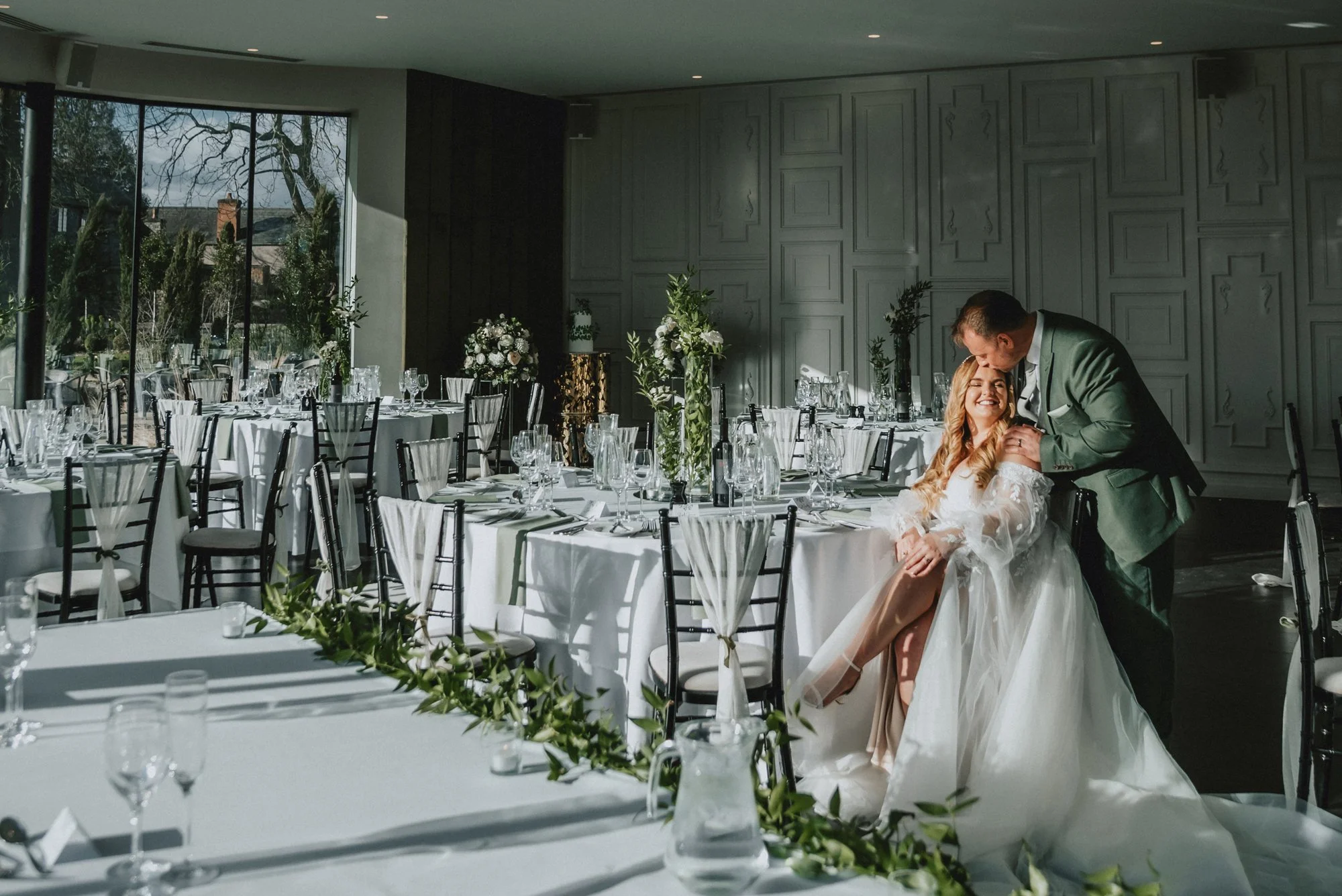 A wedding reception hall with decorated tables and chairs, sunlight coming through large windows, and a couple sharing a joyful moment, with the groom kissing the bride on her forehead.