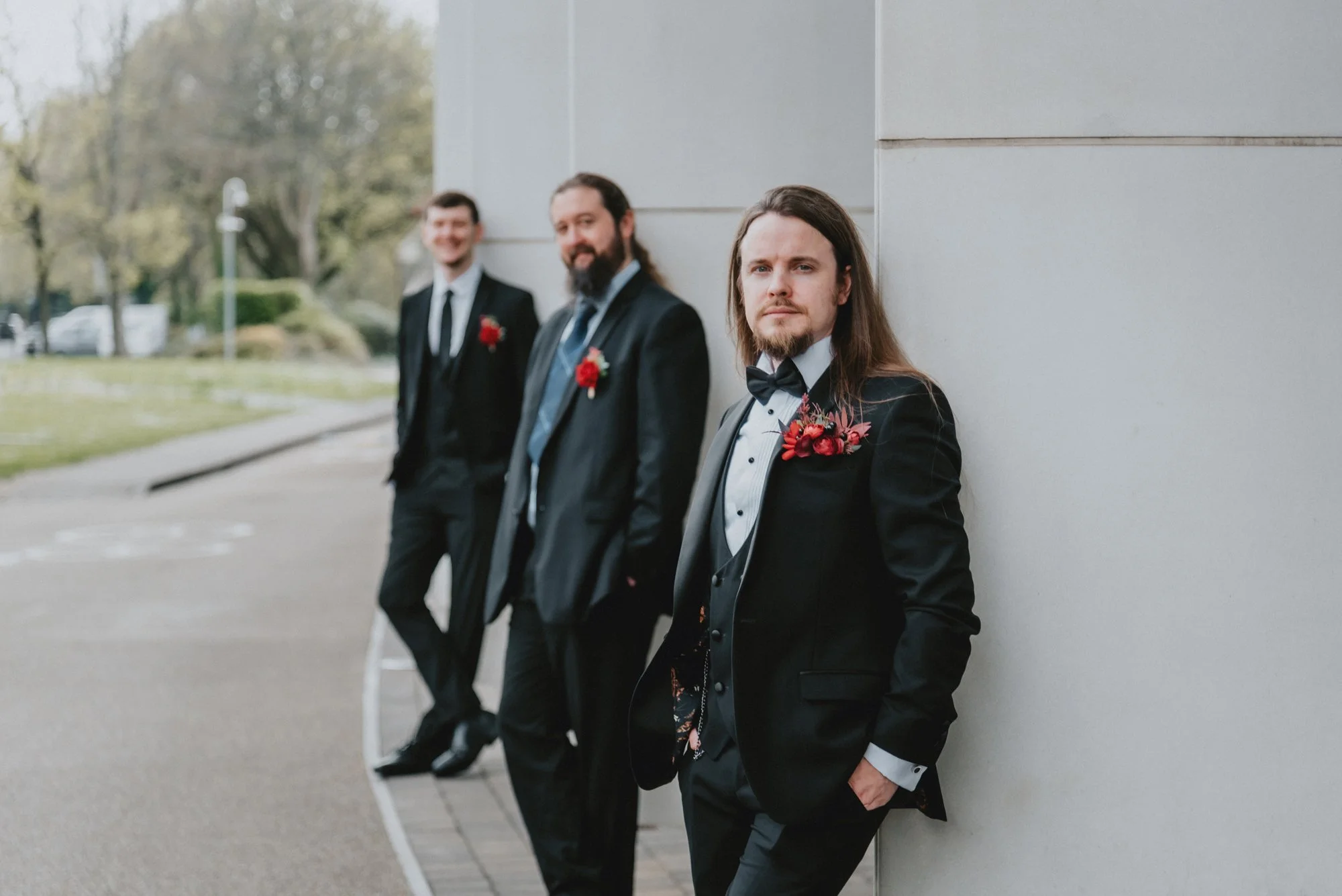 Four men in suits with red boutonnières standing against a gray building wall outdoors.