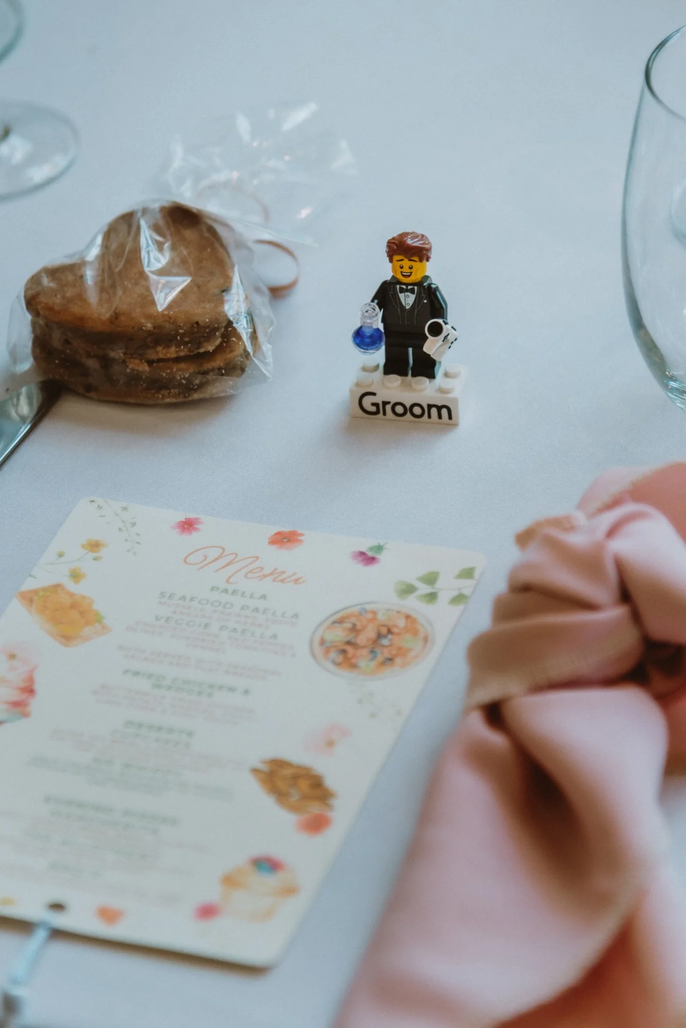 A white table with a Lego groom figure, a packaged cookie, a floral menu, and a pink scrunchie on a hand.