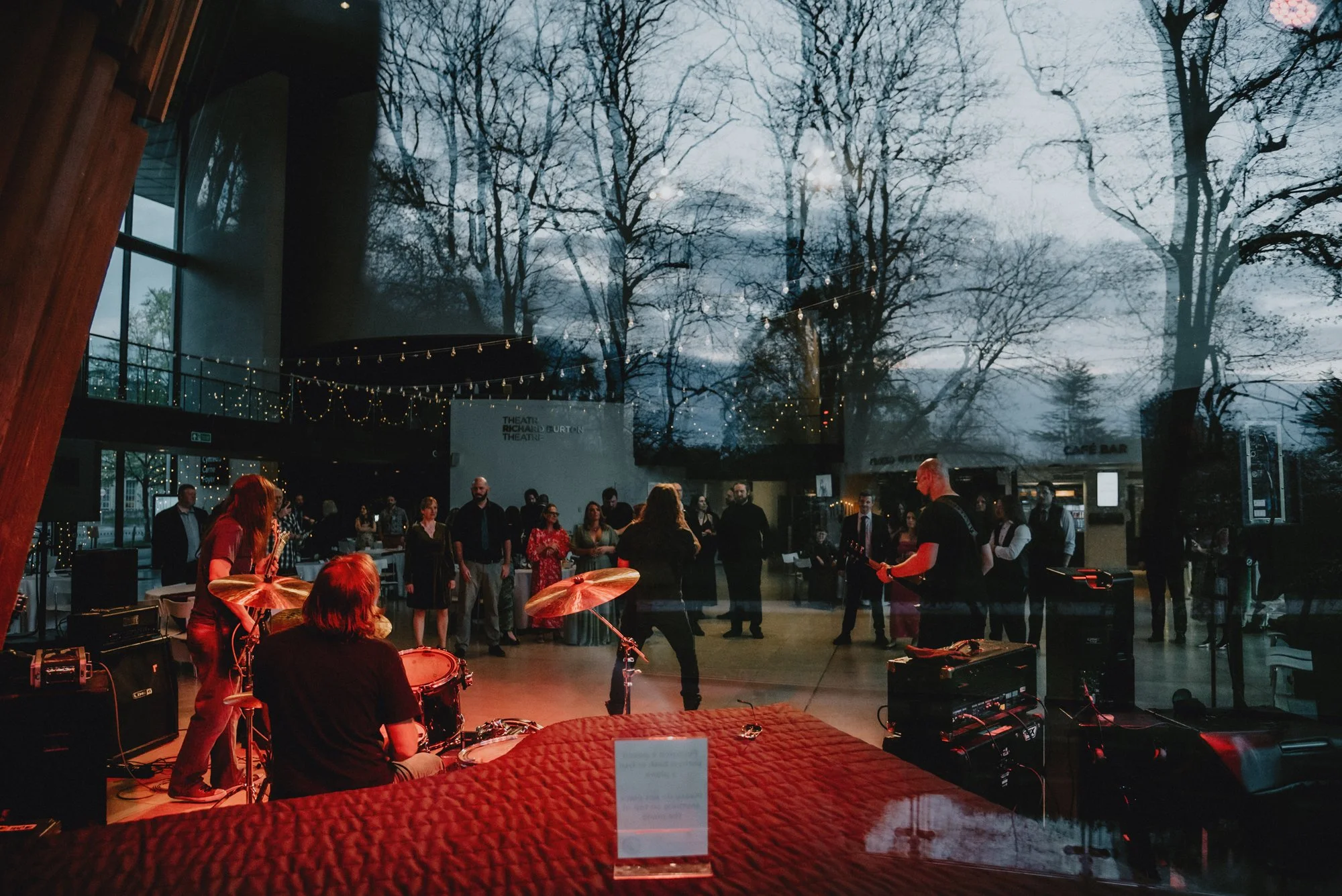 A band performs on stage at an indoor concert, viewed through a large glass window. The audience is outside, gathered around, with trees visible against a dusk sky. String lights hang inside, and a cafe bar can be seen in the background.