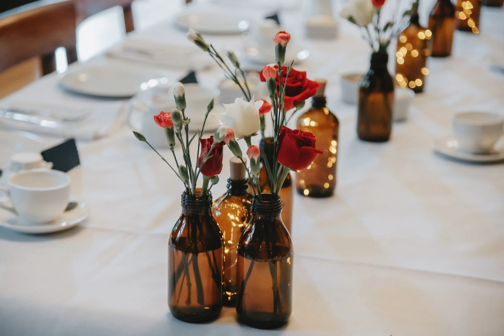 Table decorated with amber glass bottles holding red and white flowers, set with white plates, cups, and napkins.