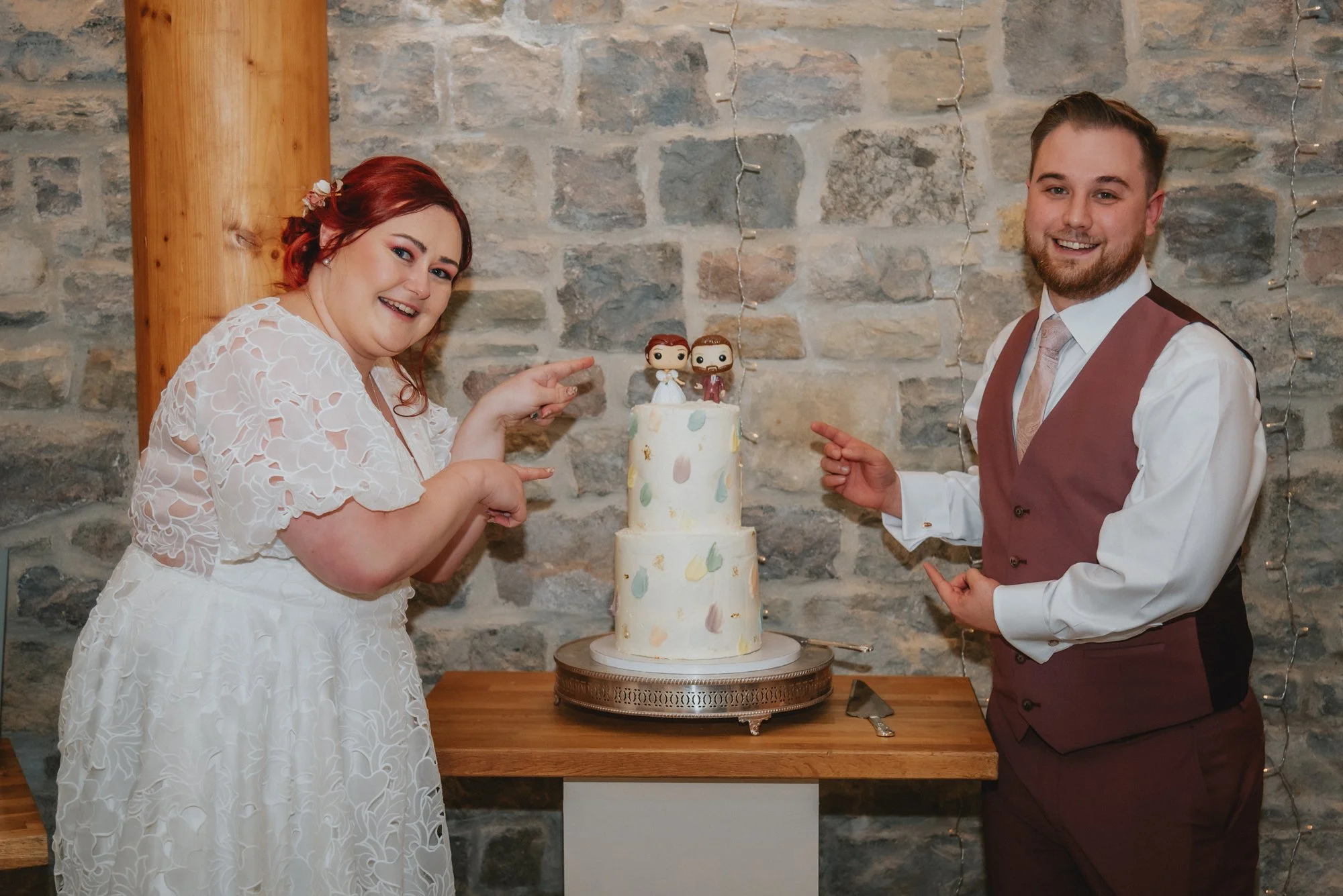 A bride and groom smiling and pointing at a wedding cake with figurines on top, standing in front of a stone wall decorated with string lights.