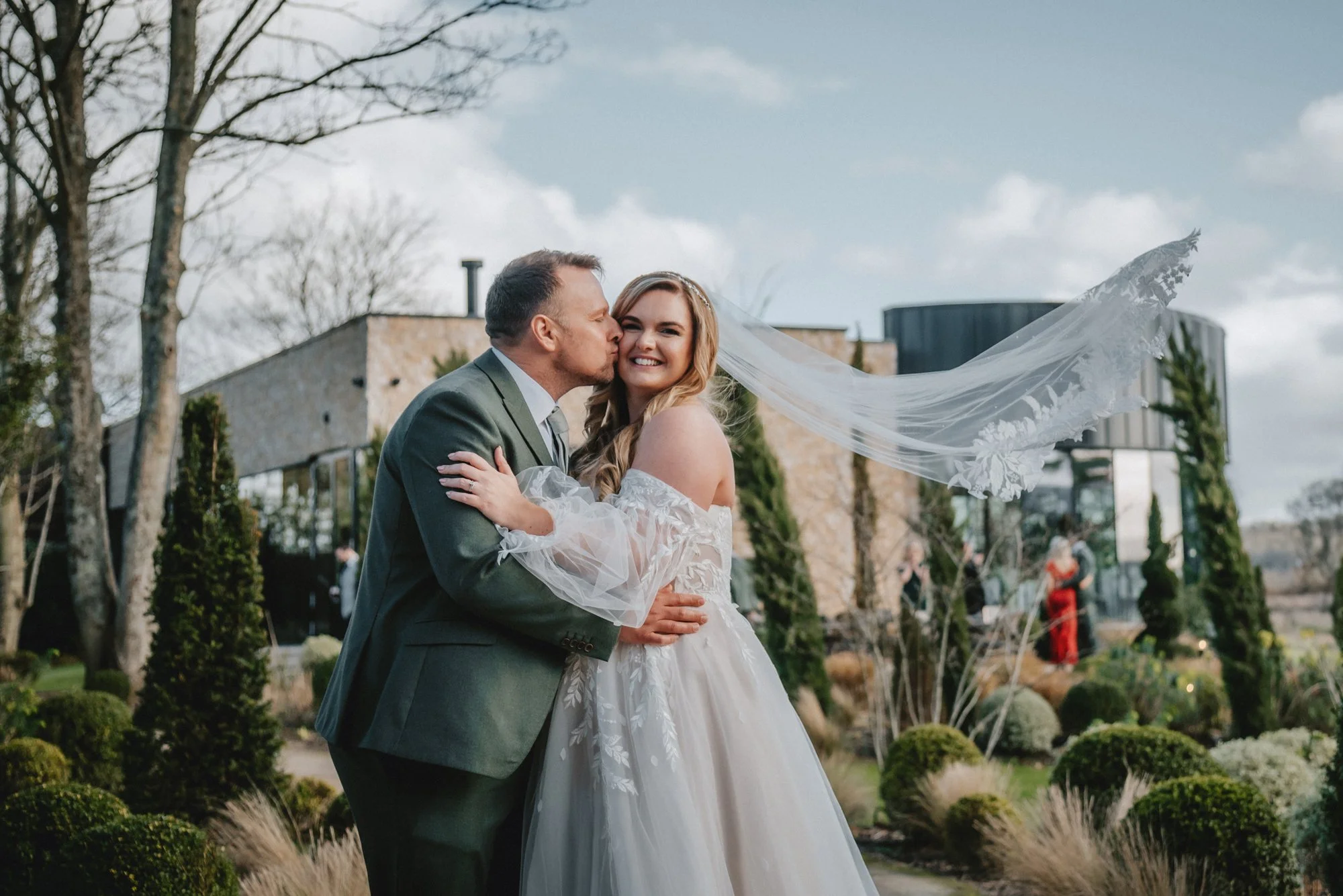 A newlywed couple embraces outdoors on their wedding day; the groom kisses the bride on the cheek as she smiles. The bride wears a white wedding gown with a veil flowing in the wind, and the groom is in a gray suit. In the background, there are trees