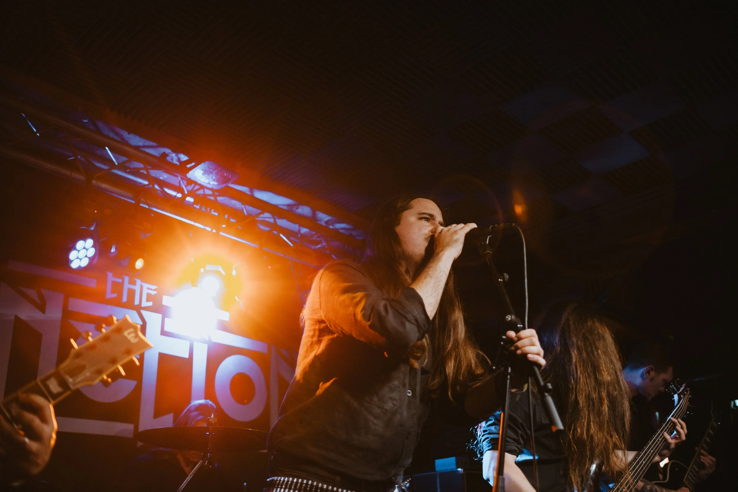 Female singer performing on stage with band members, colorful stage lighting and a backdrop with the band name.