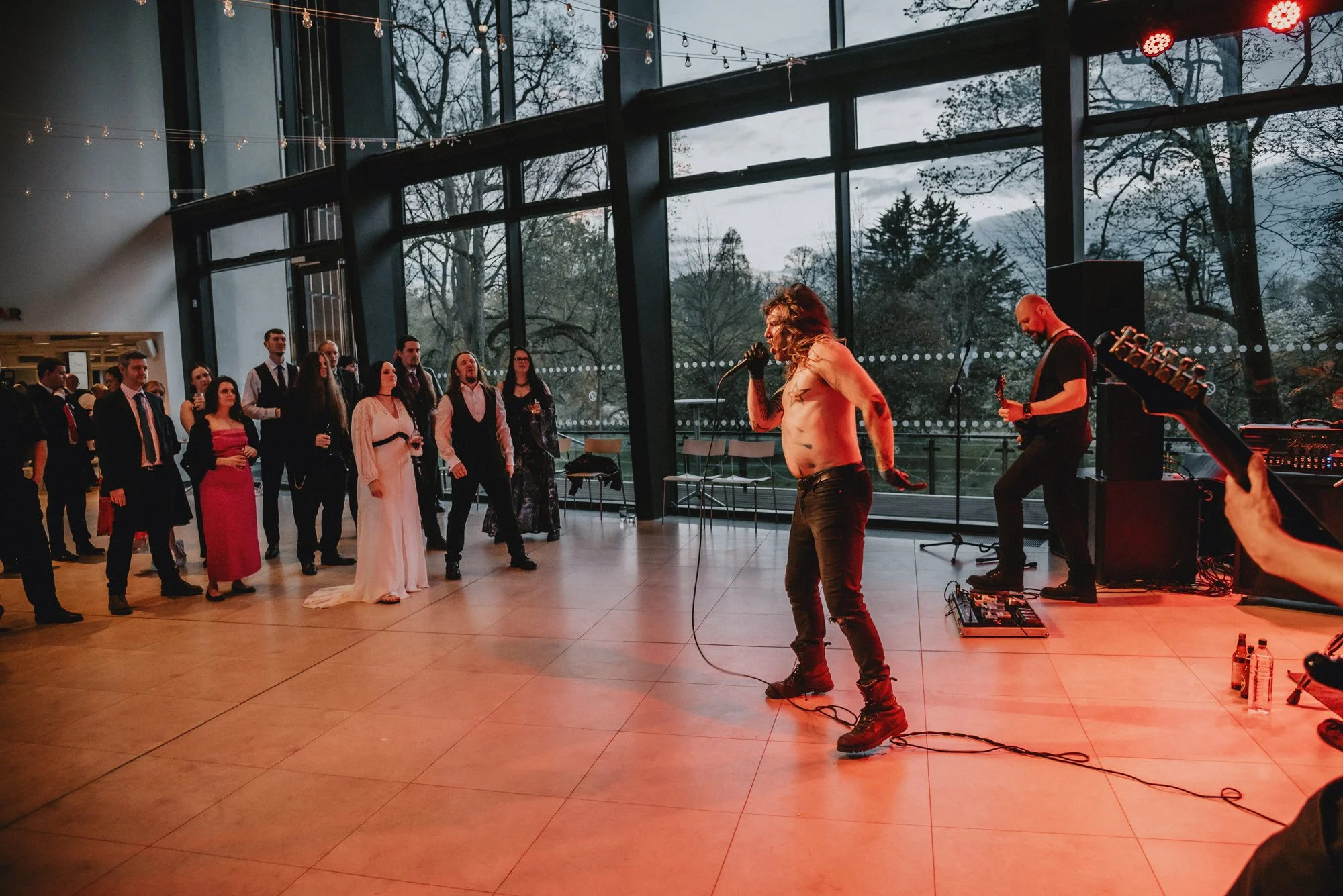 A shirtless singer with long hair, tattoos, and black pants performs with a mic at a modern event space with large windows showing an outdoor view. Several guests in formal and semi-formal attire dance and watch in the background. A guitarist is play