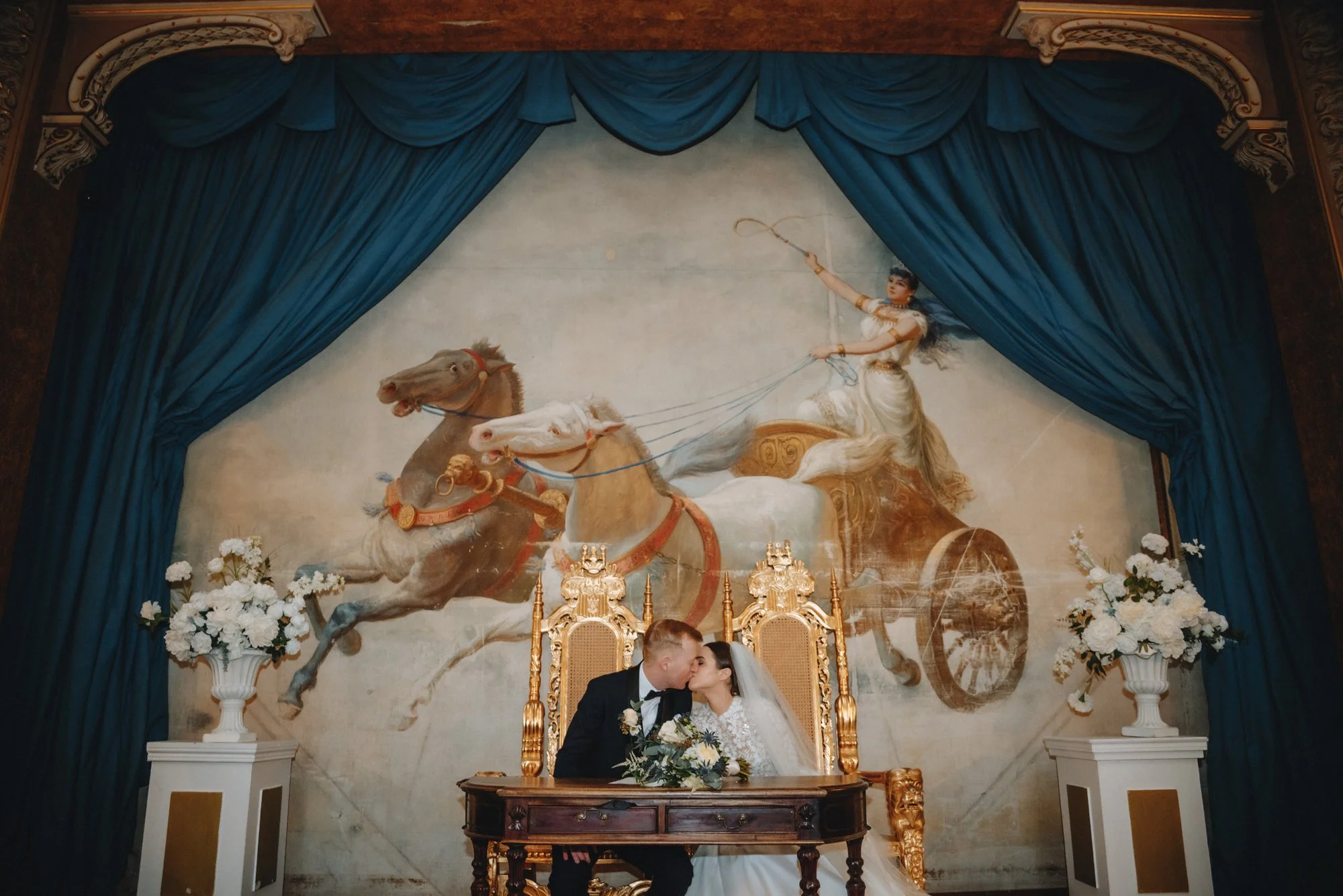 A newlywed couple kissing at a wedding ceremony, seated at a ornate wooden table with a large mural of a chariot drawn by horses behind them, framed by rich blue drapes and white floral arrangements.