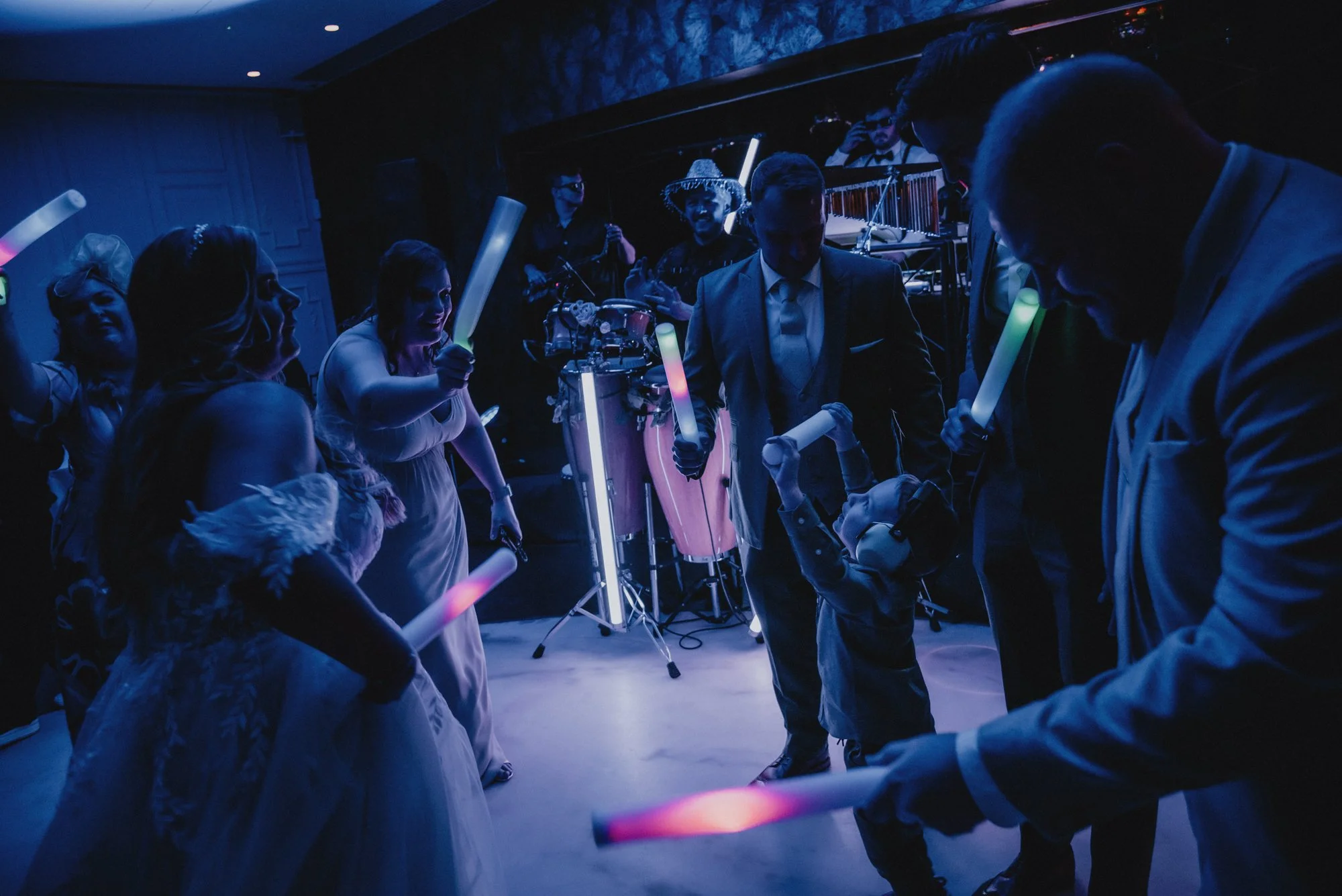 Guests dancing and celebrating at a wedding reception with glow sticks, a live band playing in the background, and dim lighting.