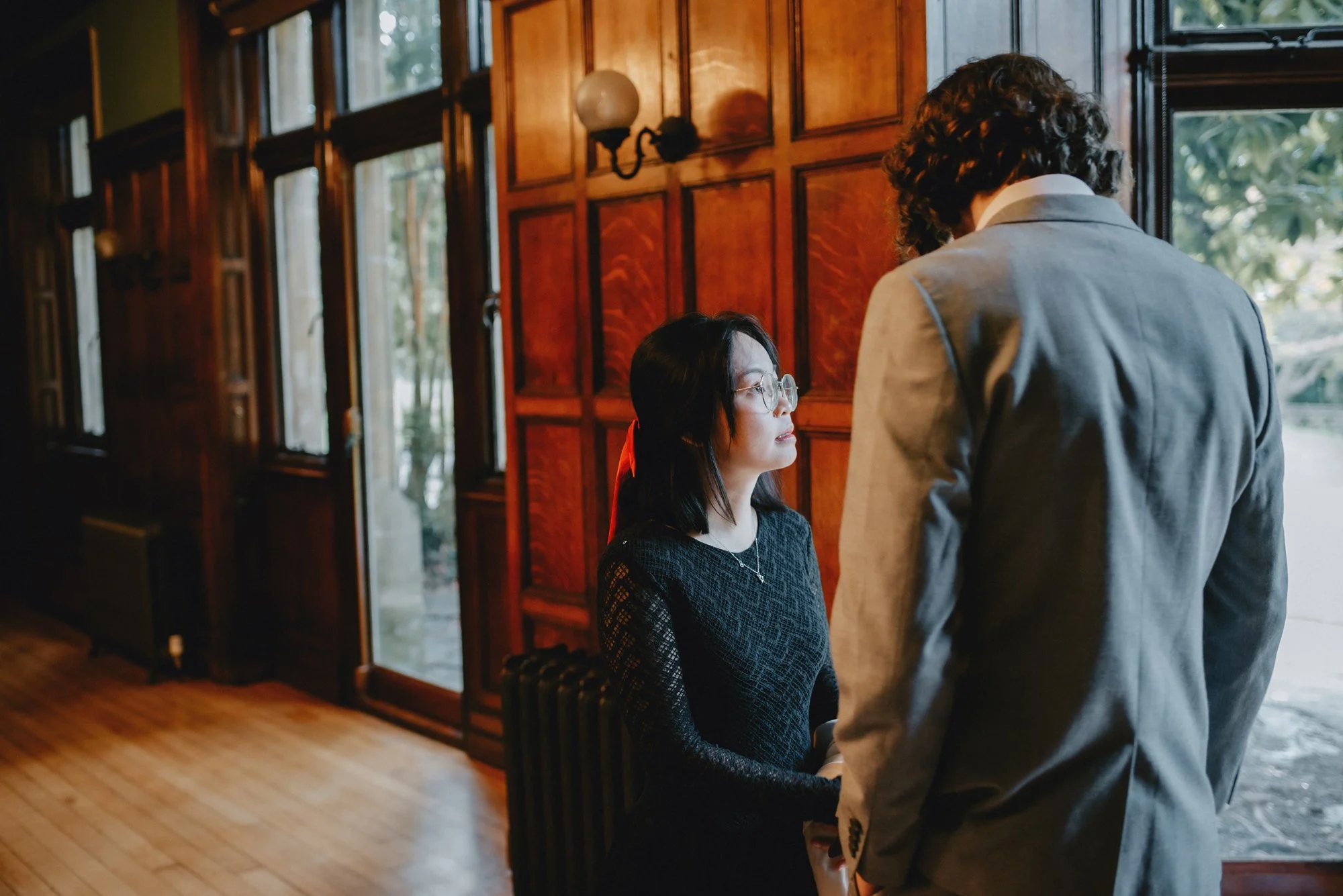 A woman with glasses in a black dress and a man in a gray suit stand close together inside a room with dark wood paneled walls and large windows, engaged in conversation.