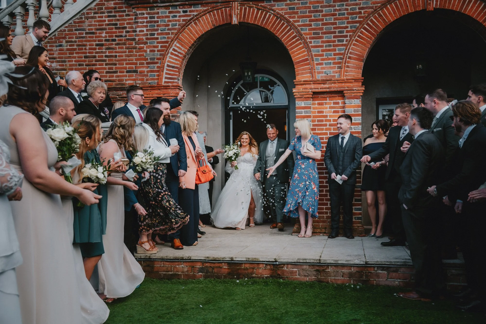 A bride and her parents walking out of a brick building, surrounded by friends and family celebrating with confetti at a wedding.