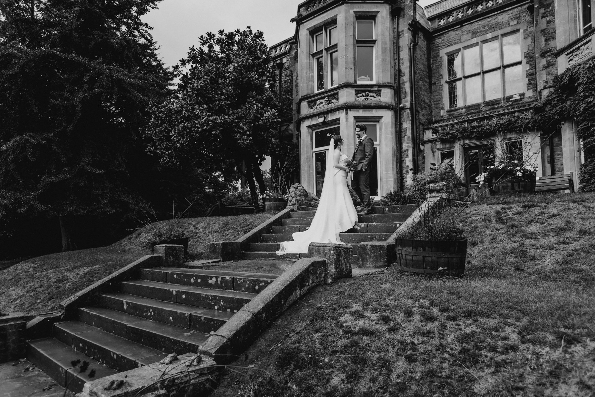 A bride and groom standing on outdoor stairs in front of an old stone house, holding hands and facing each other, during a wedding photoshoot.